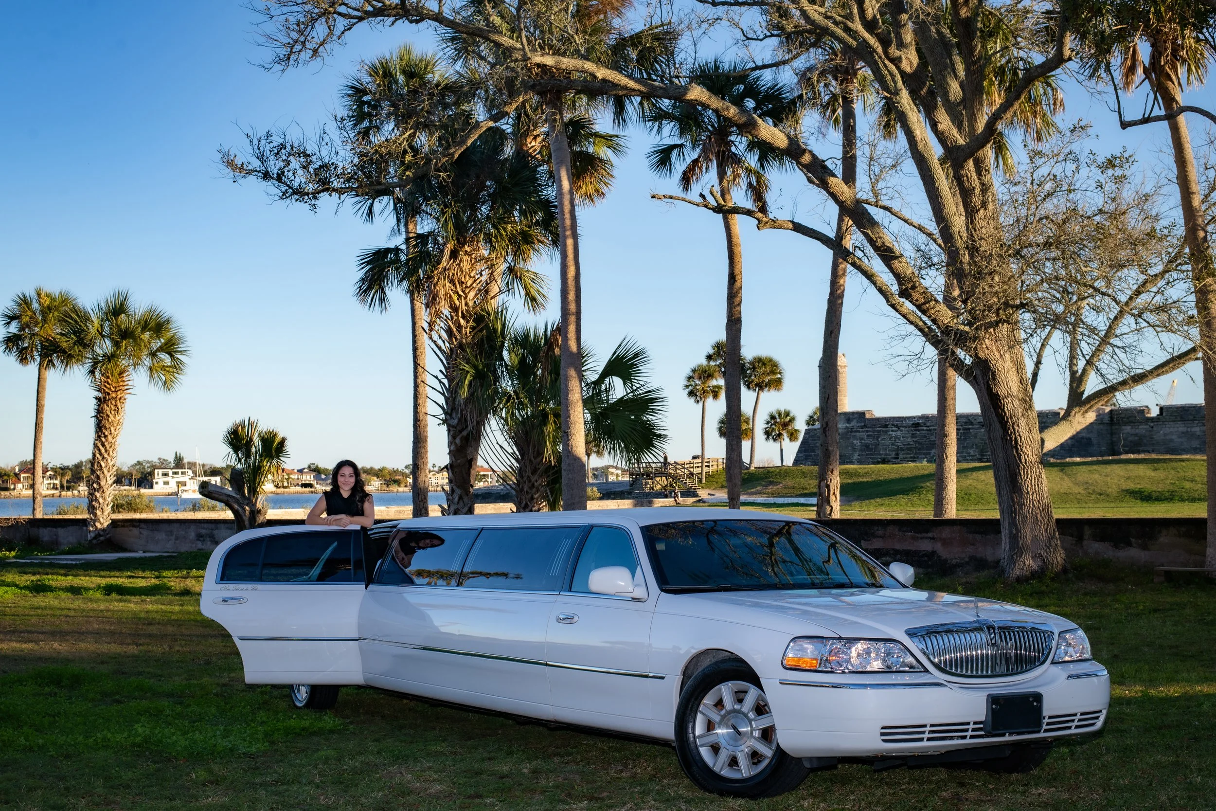 A woman standing next to a white limousine parked on a grassy area with palm trees and a river in the background, under a partly cloudy sky.