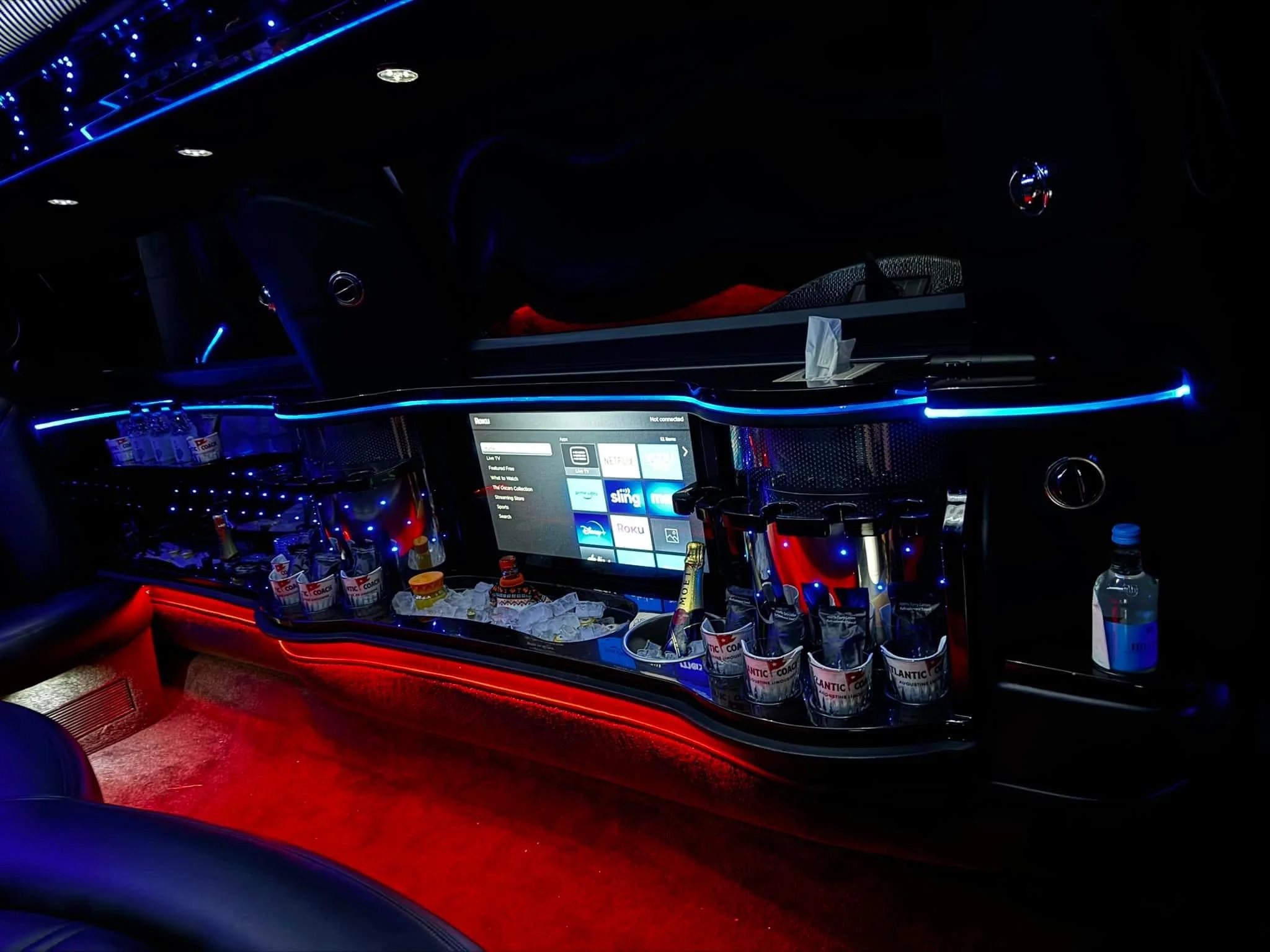 Inside a limousine featuring a bar with alcoholic beverages, a lighted infotainment screen displaying streaming options, and LED lighting accents in red and blue.
