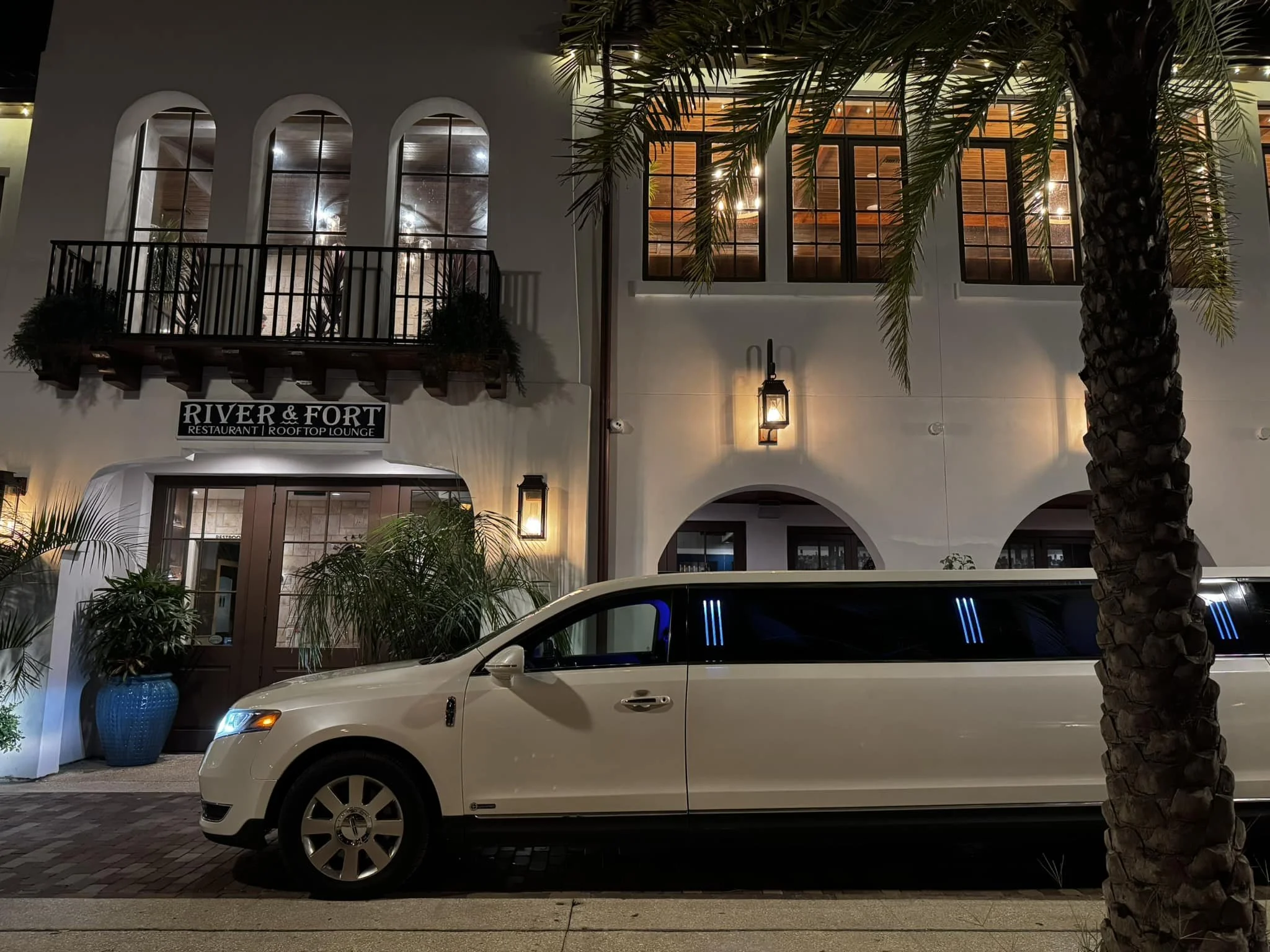 White limousine parked outside a restaurant and rooftop lounge named River & Fort at night, with palm trees and building lights.