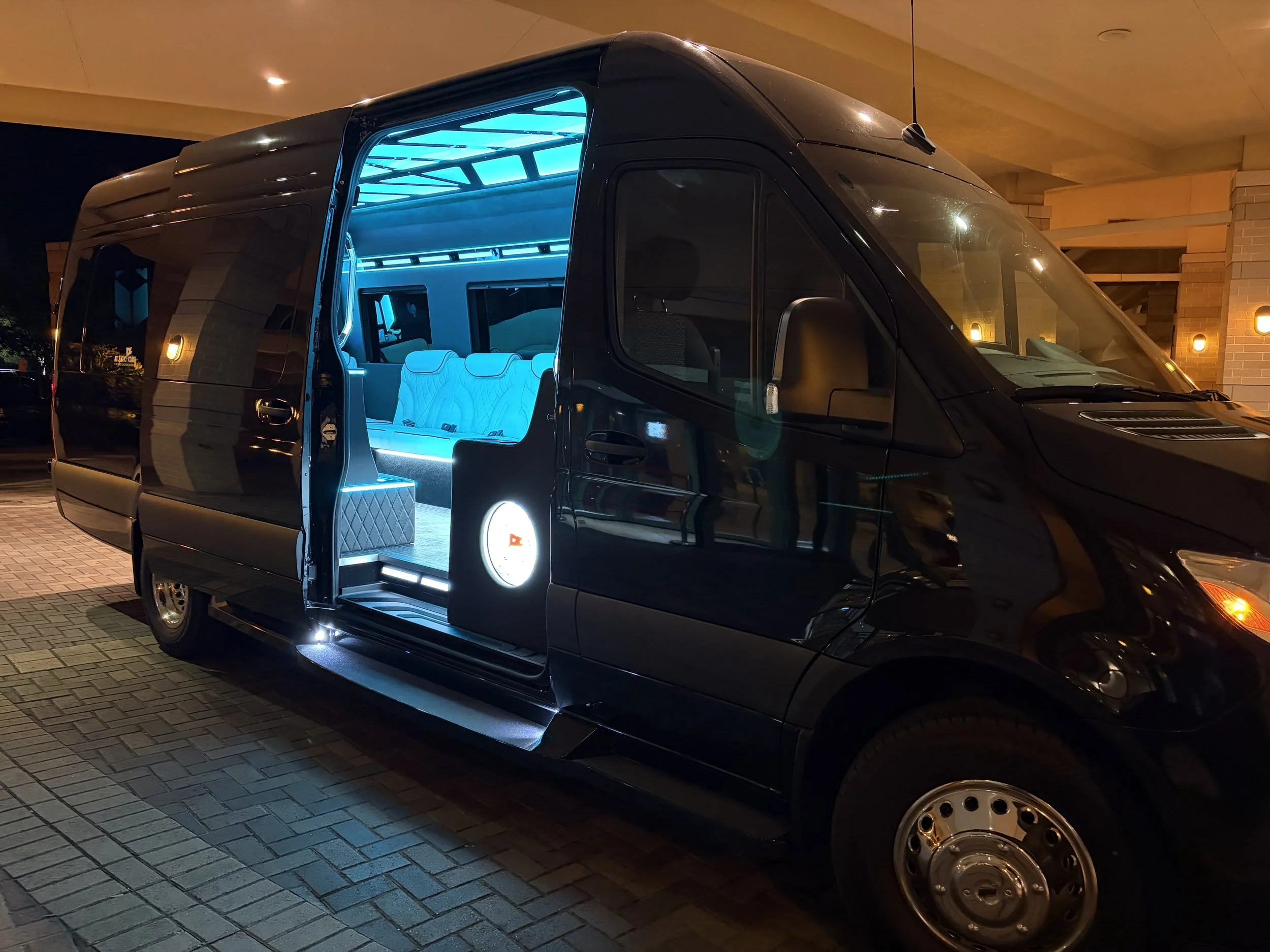 A black camper van at night with doors open, revealing illuminated interior with blue lighting and white quilted seating.