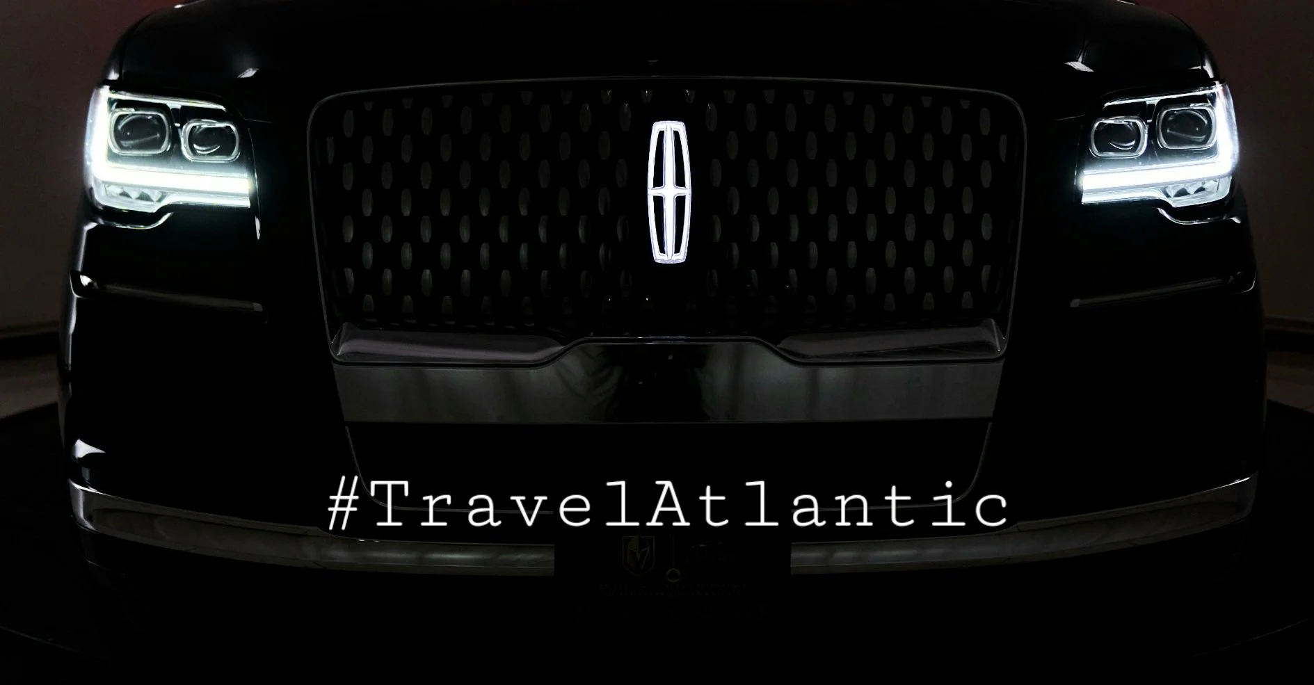Front view of a black luxury car with illuminated headlights and a prominent grille, featuring the Lincoln emblem, with the hashtag #TravelAtlantic overlayed.