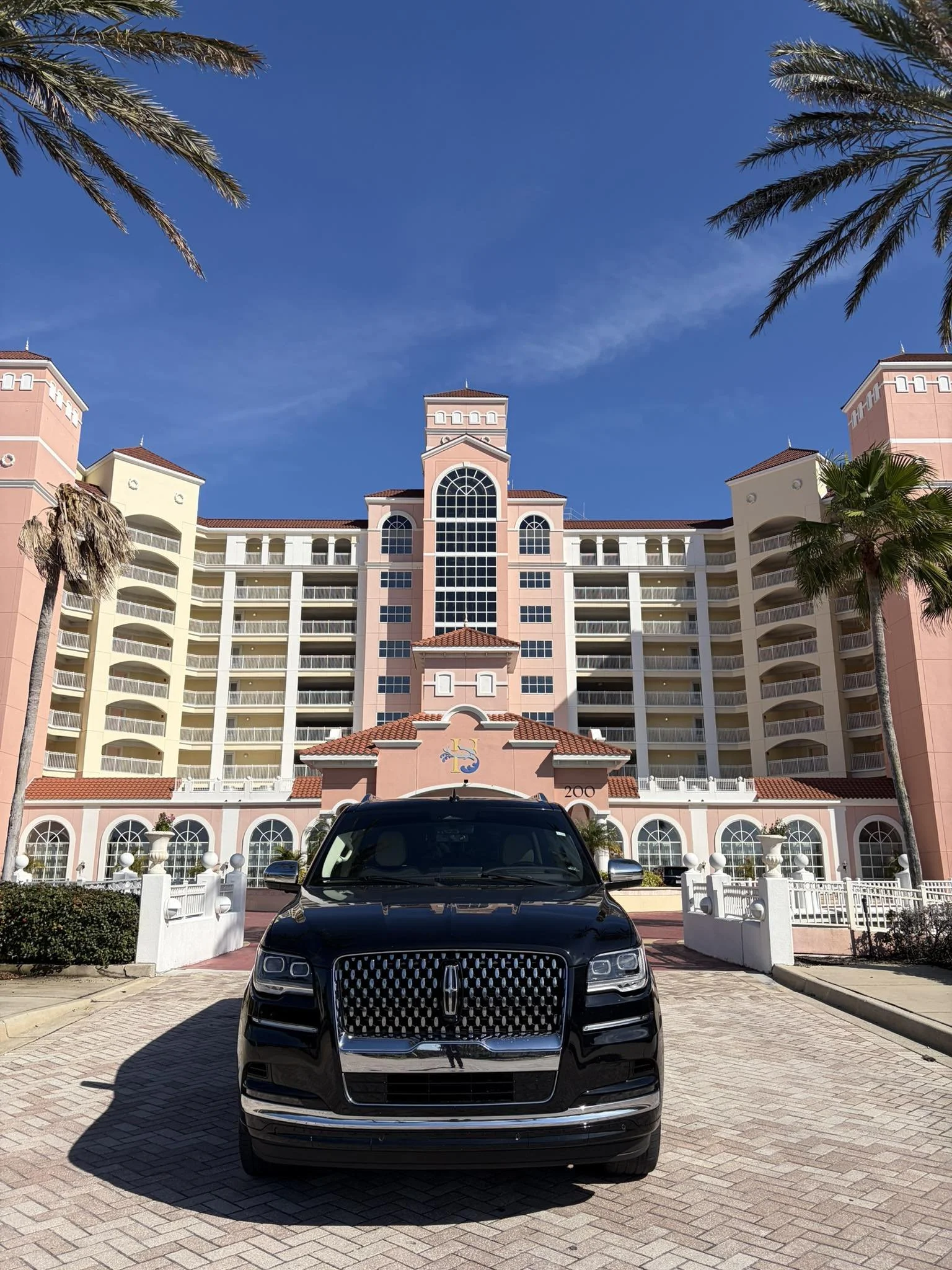 Black Lincoln SUV parked in front of a pink and white seaside hotel with palm trees under a blue sky.