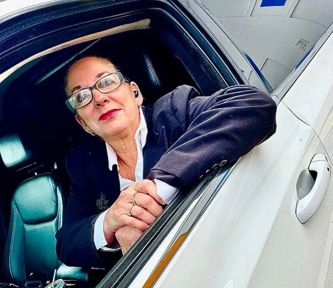 A woman in glasses and business attire leaning out of a car window, shaking hands with someone off-camera.