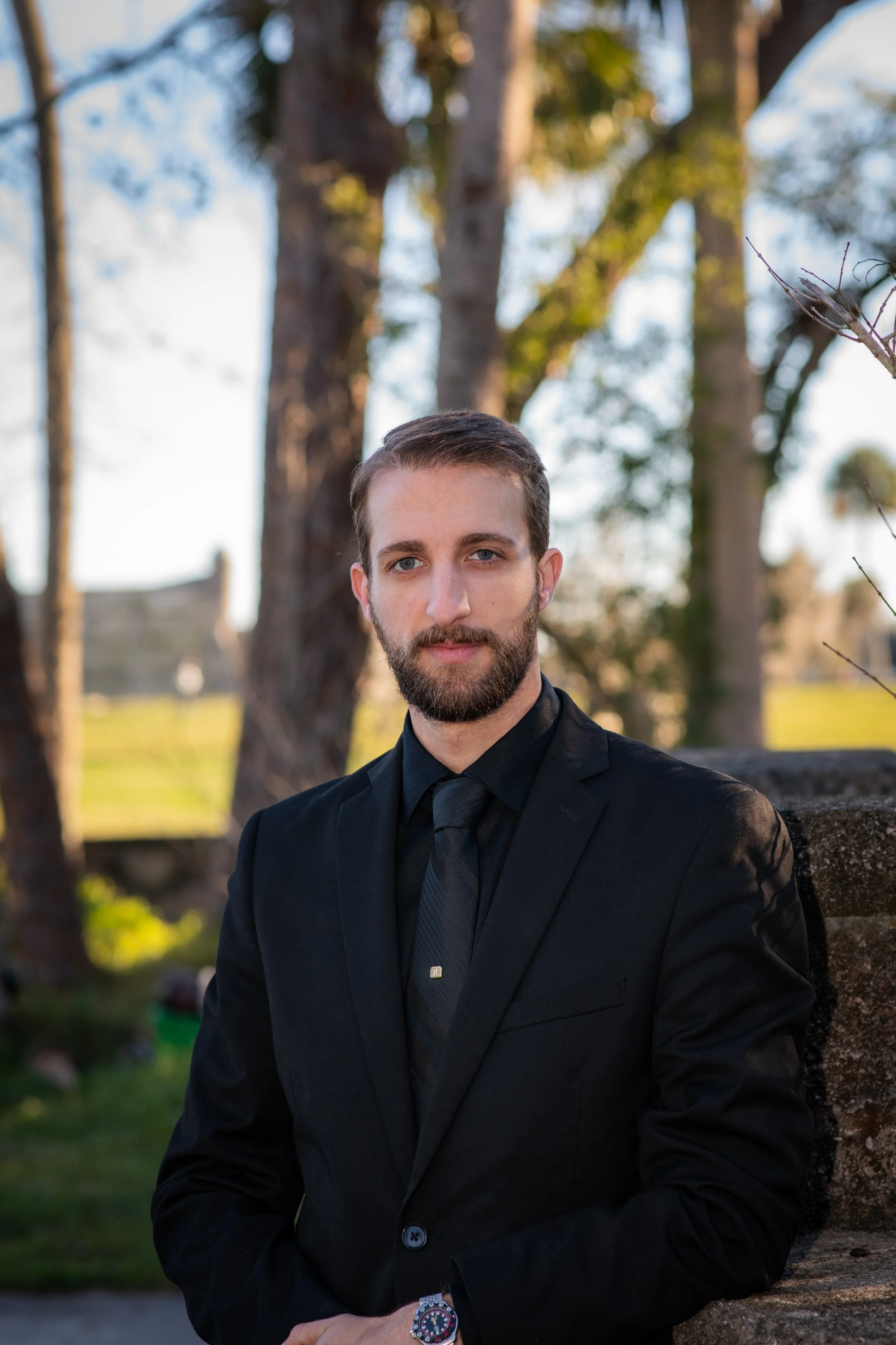 A man in a black suit and tie standing outdoors with trees and a stone wall in the background.