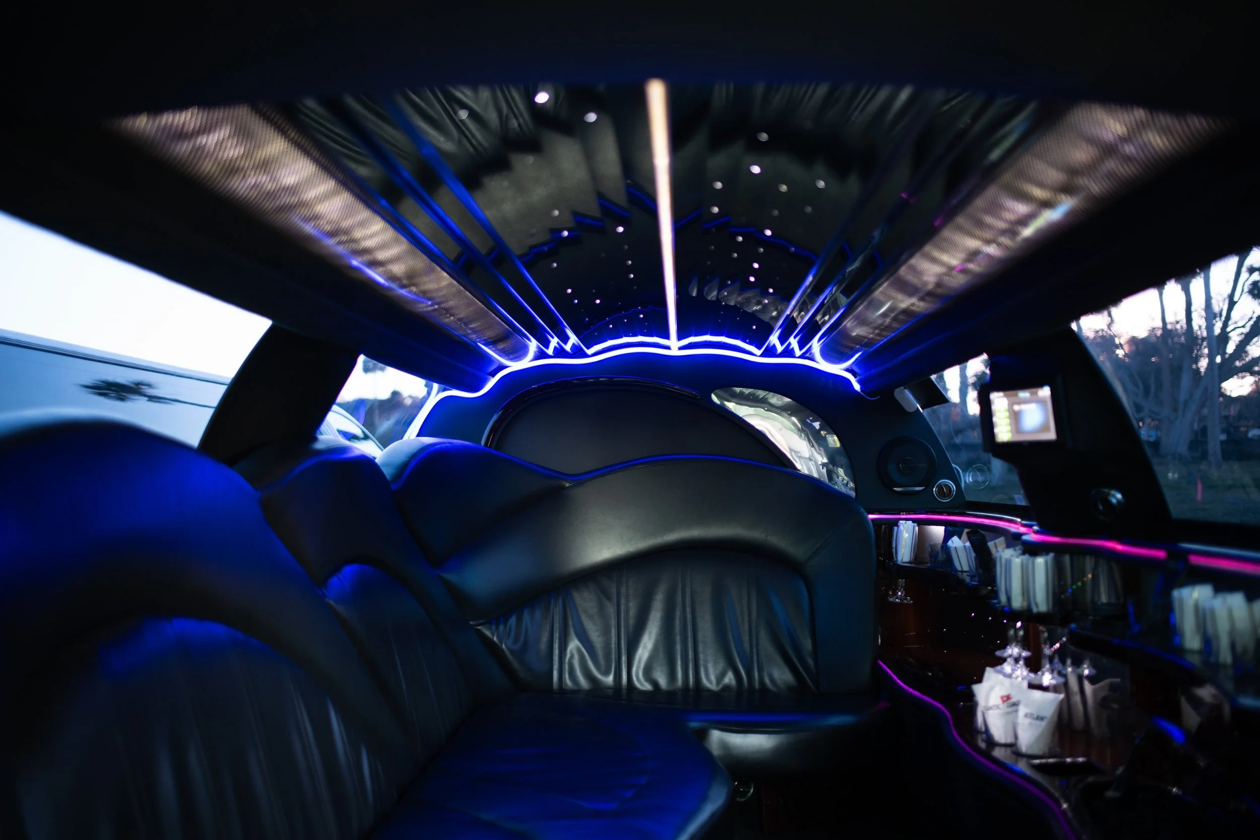 Inside a limousine with black leather seats, illuminated by neon purple and blue lights, with a bar area holding glasses and napkins, and a mirror on the ceiling reflecting the lights.