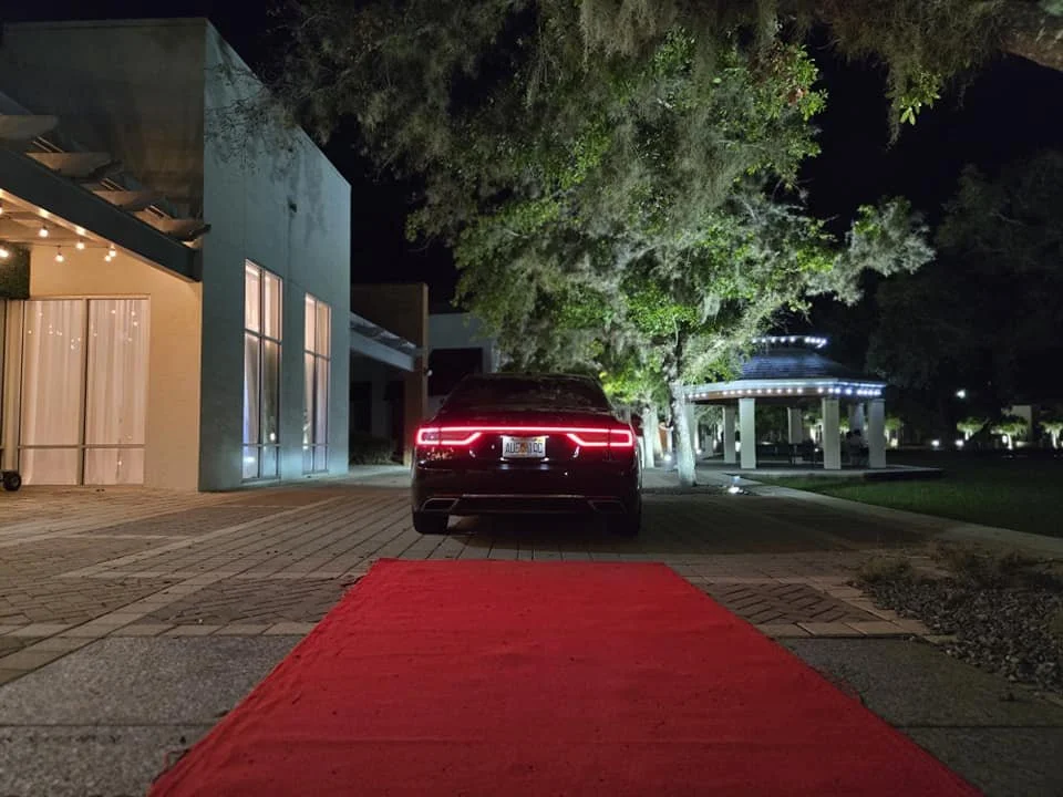 A black car with illuminated taillights parked on a driveway next to a building at night, with a red carpet leading up to the car, trees and a lit pavilion in the background.