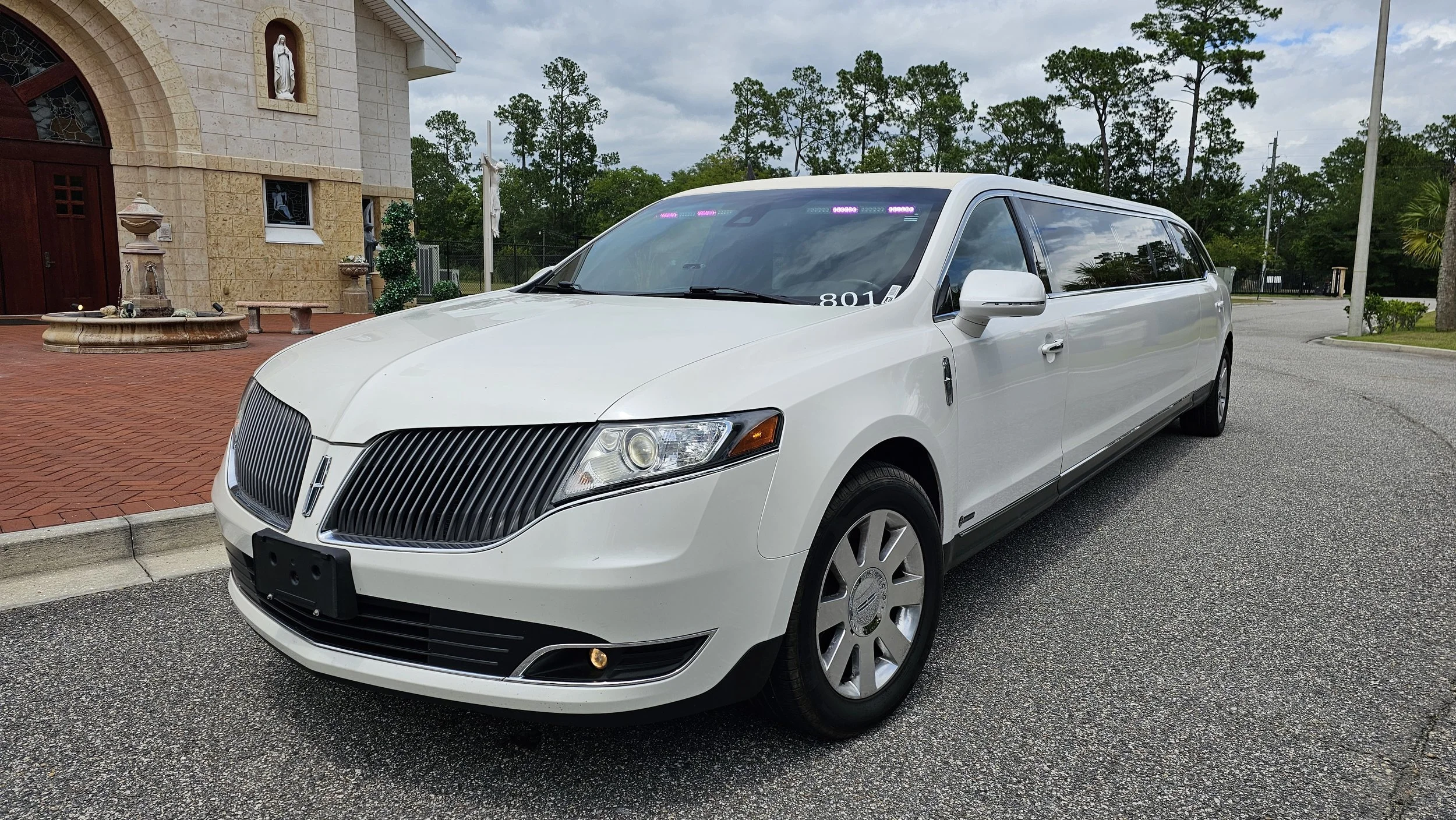 A white limousine parked on a street in front of a church with a fountain.