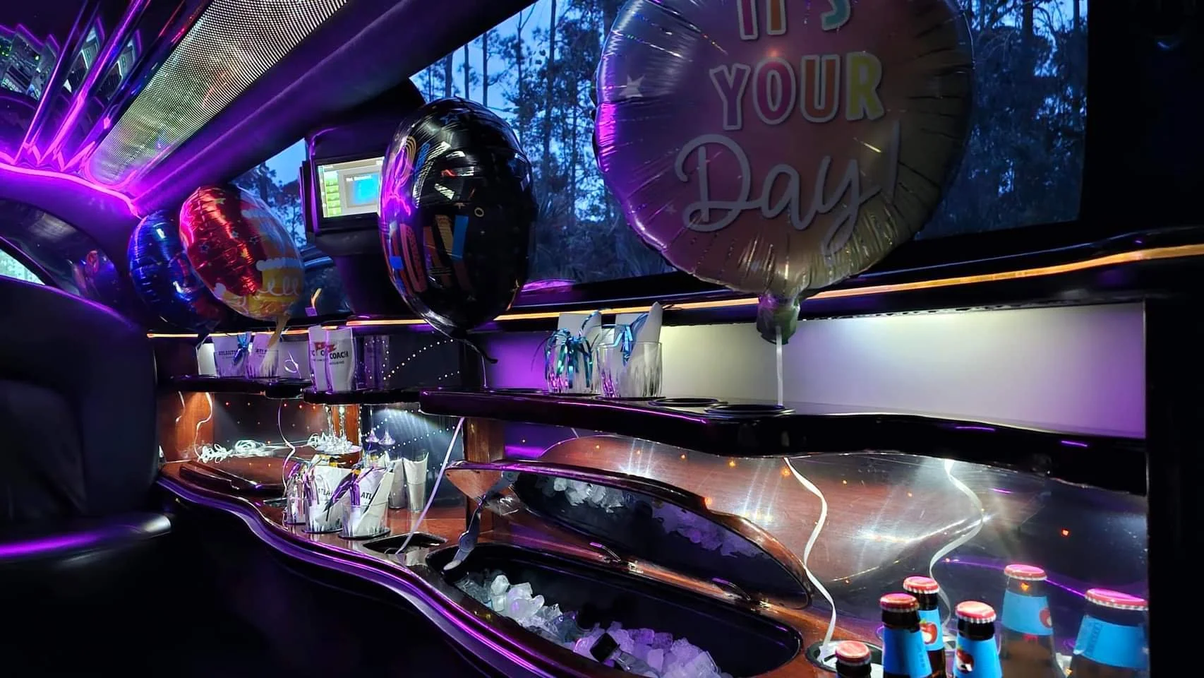 Decorated bus interior with purple neon lights, black balloons with colorful messages including "It's Your Day," and drink bottles. The shelves have party supplies, and the scene appears to be set for a celebration.