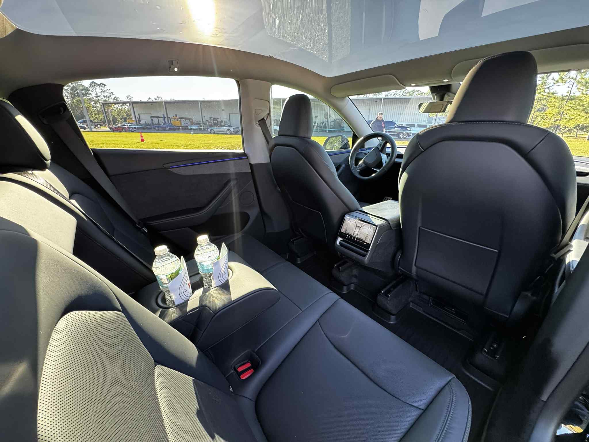Interior of a modern vehicle with black leather seats, two water bottles in a cup holder on the rear seat, and a view of an outside parking area with buildings and trees.