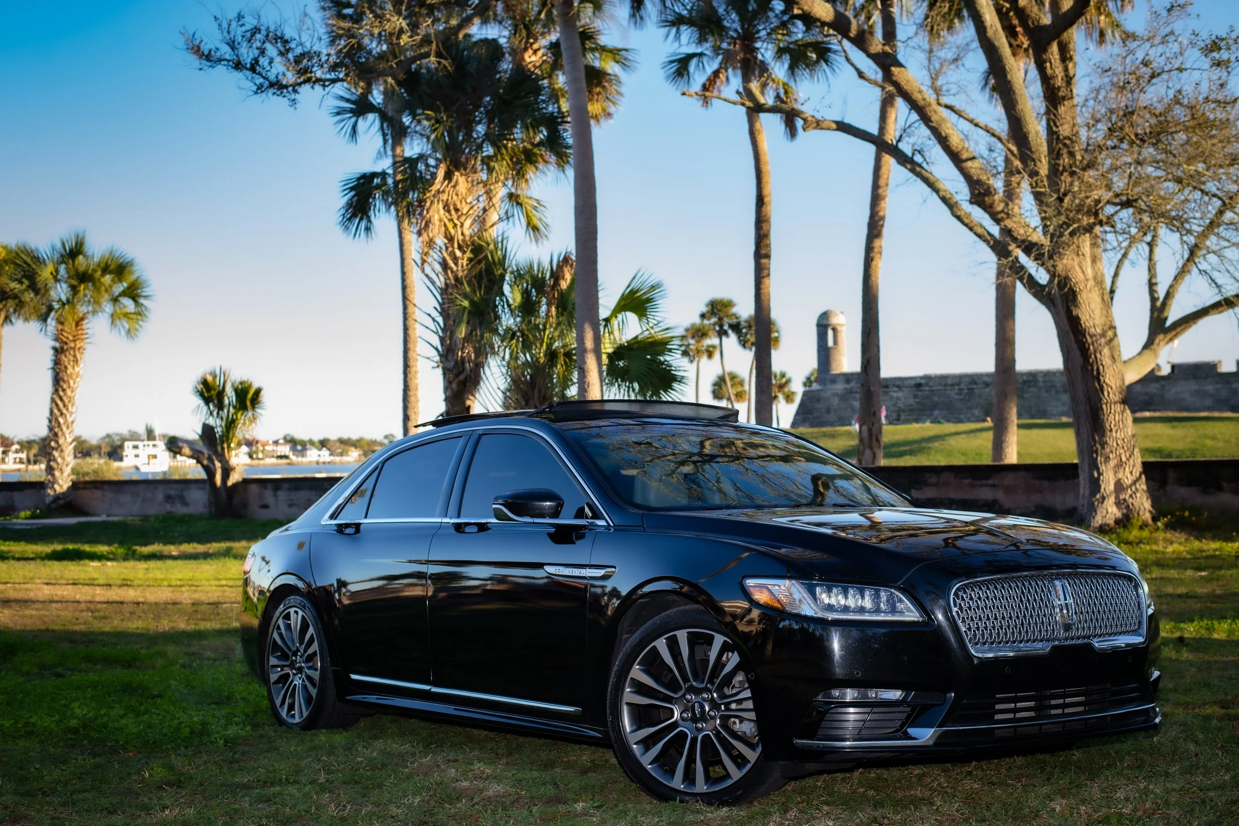 Black luxury sedan parked on grass with palm trees and historic fort in the background under a clear sky.
