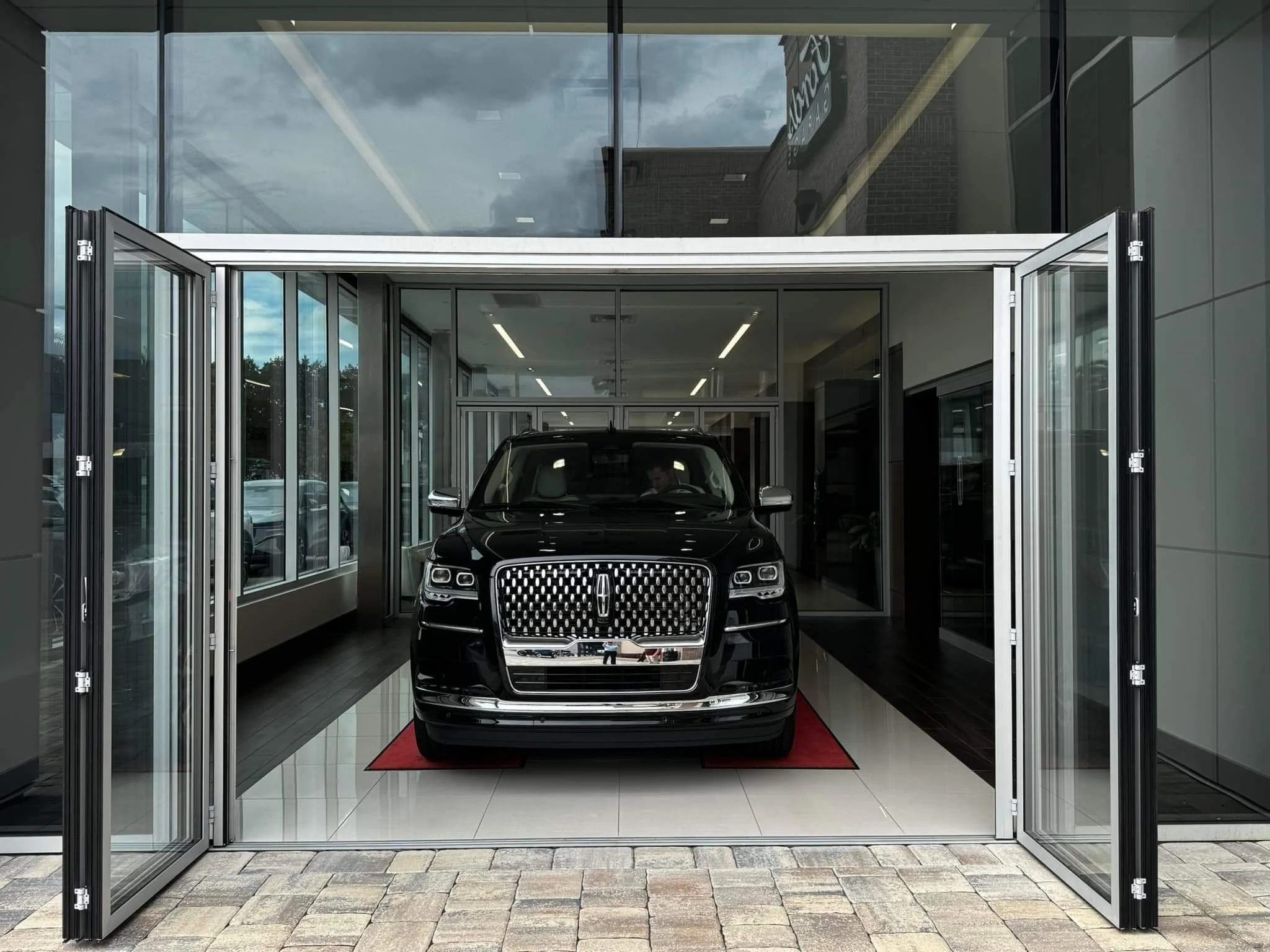 Black luxury SUV inside a modern car dealership with glass walls and open glass doors, displayed on a white tiled floor with a red carpet under the vehicle, and a cloudy sky reflected in the glass ceiling.