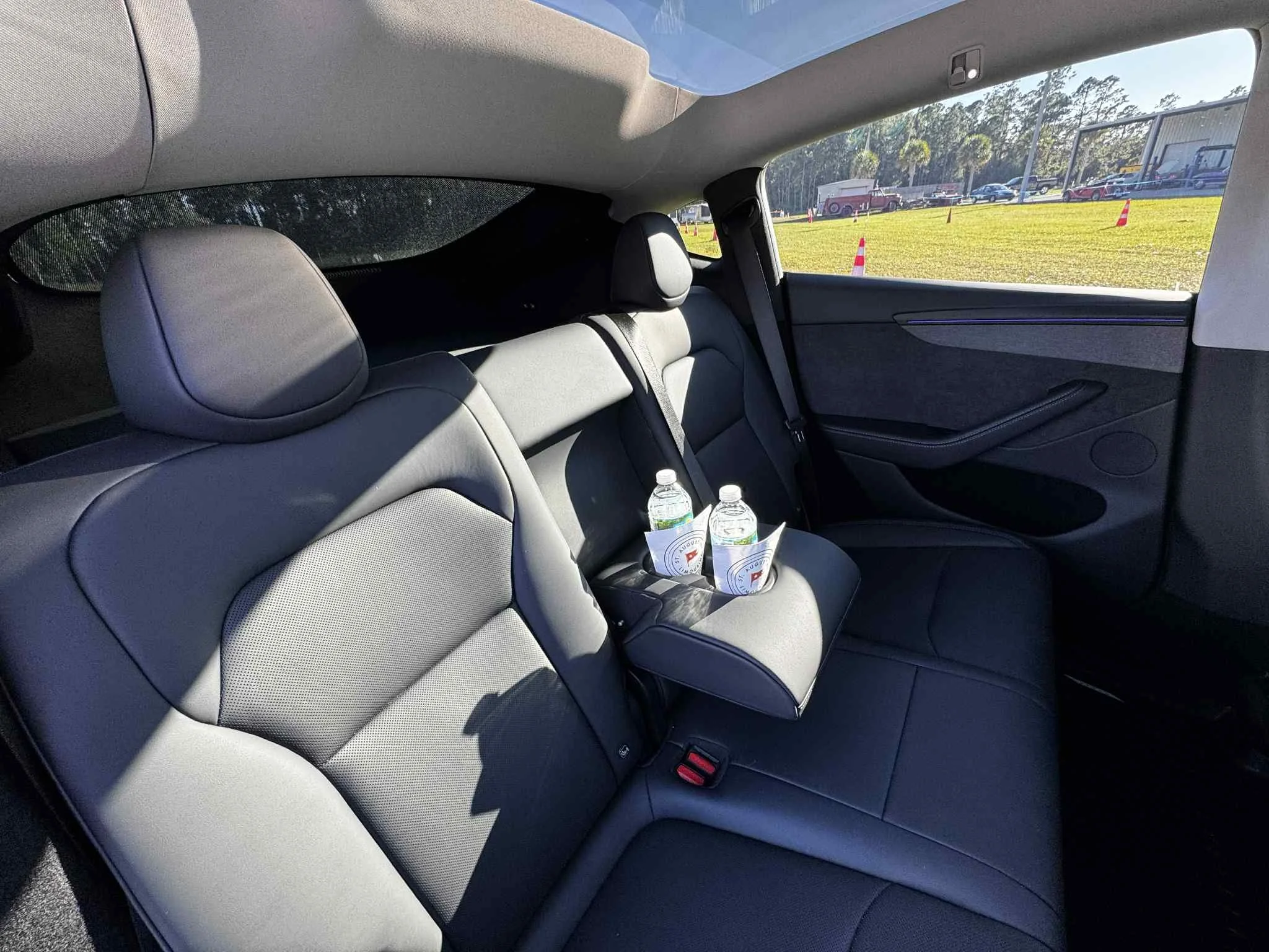 Back seat of a car with two water bottles and a cup holder with a logo, outside view of a parking lot with cars and trees.