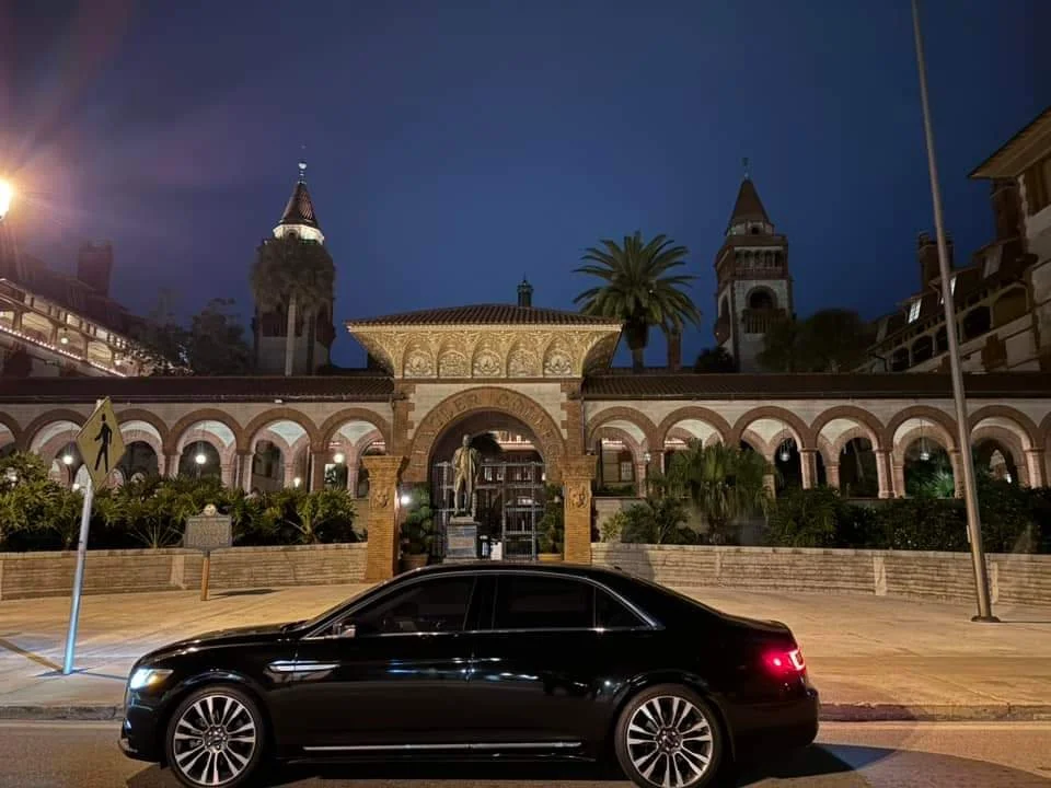 A black car parked in front of a historic building with arched windows and towers, at night with streetlights illuminating the scene.