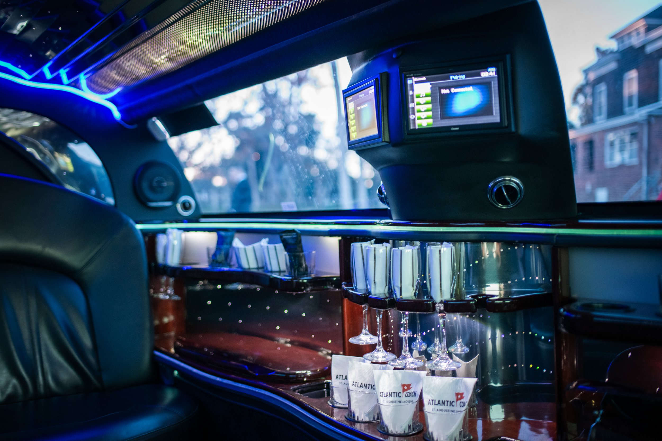Interior of a luxury limousine with glassware and digital screens, and a cityscape visible through the windows.