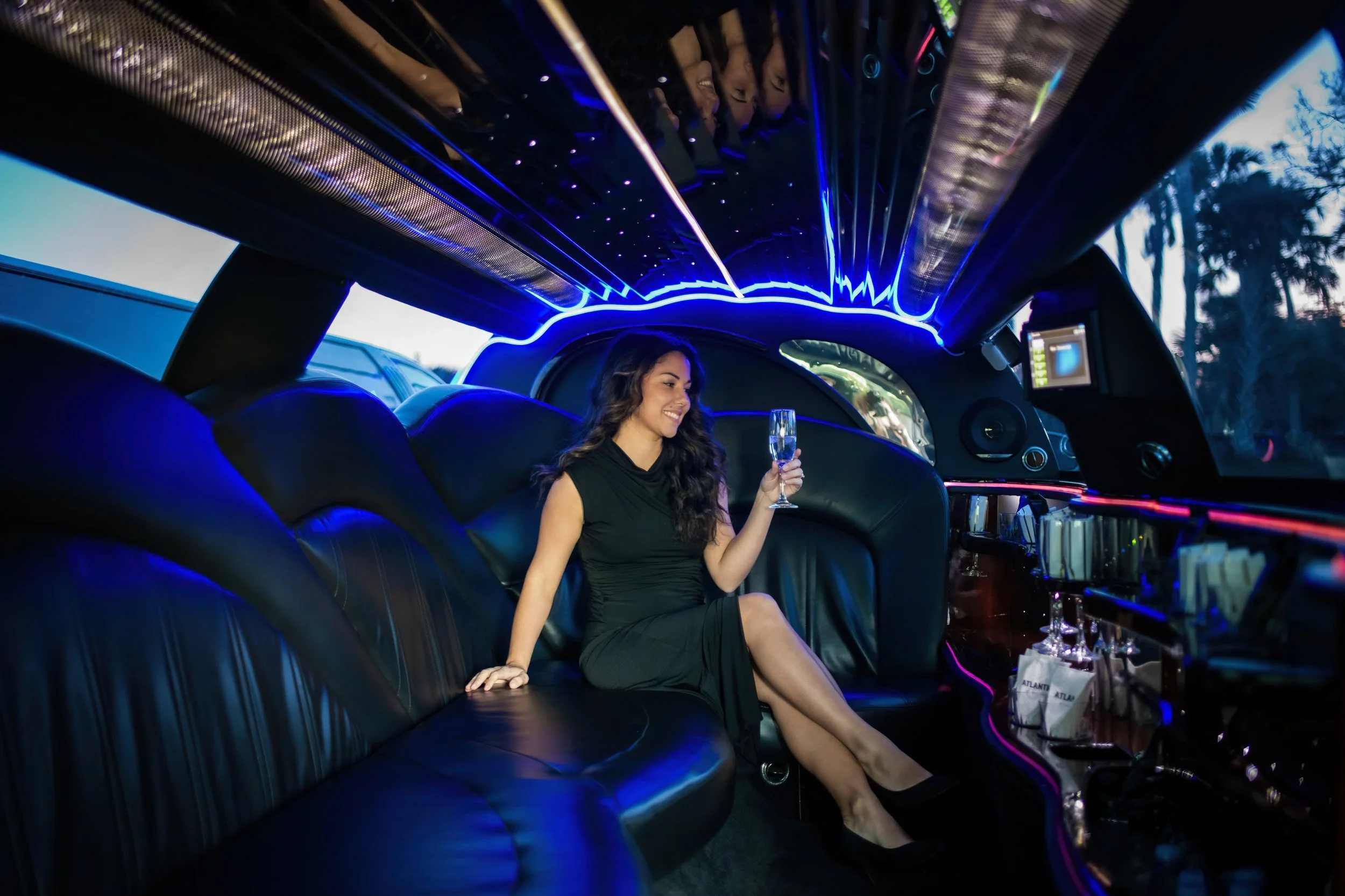 A woman in a black dress sitting inside a limousine with black leather seats, holding a glass of champagne and smiling, with blue and purple neon lighting reflecting on the ceiling and surroundings.