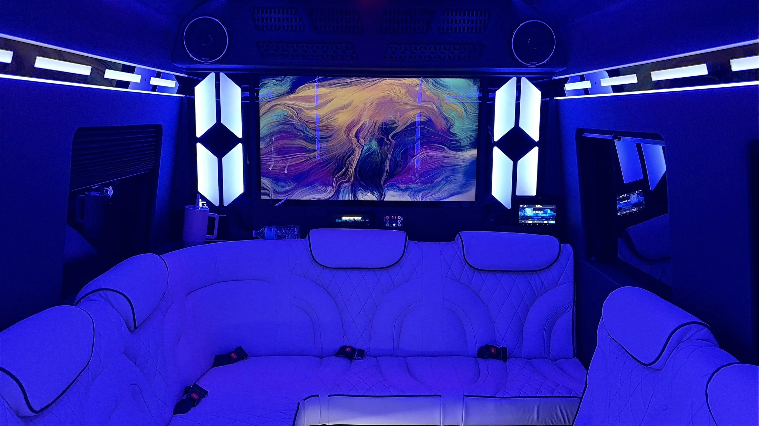 A futuristic spaceship lounge with a U-shaped white couch, illuminated by blue neon lights, a large abstract digital art display on the wall, and smaller screens on the sides.