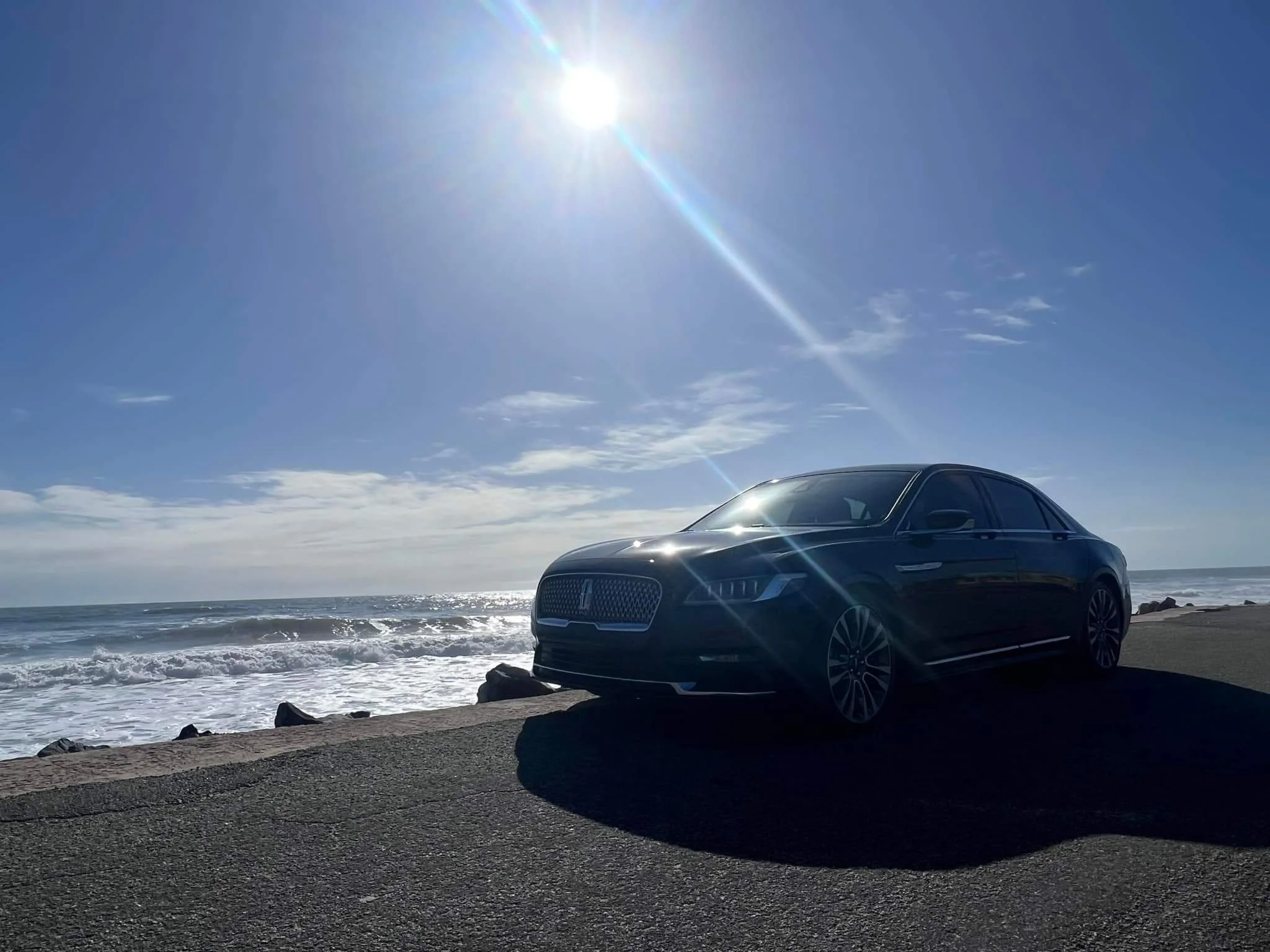 A black luxury sedan parked near the ocean under a bright sun, with waves crashing onto the shore in the background.