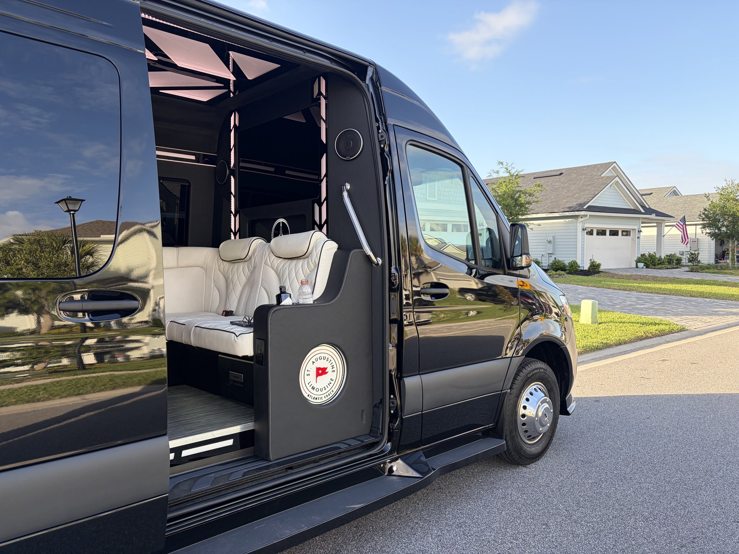 A black limousine parked in a suburban neighborhood with houses and a blue sky in the background. The limousine is designed for luxury and has seating and sound equipment inside.
