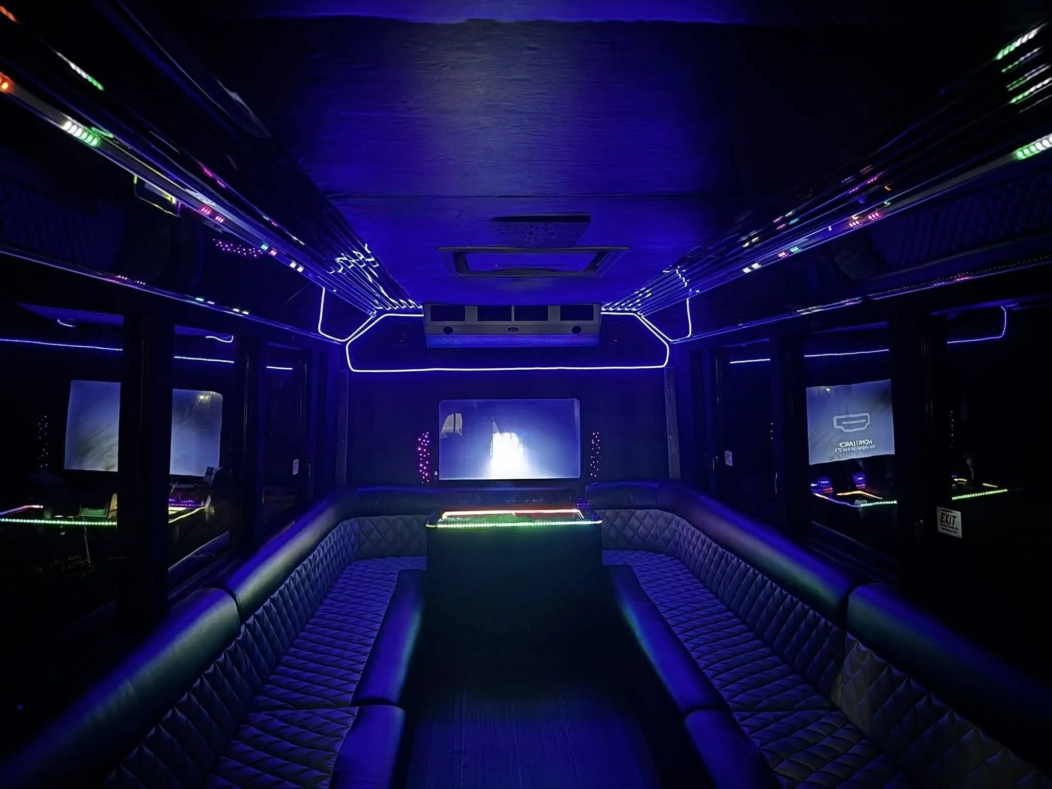 Interior of a dark, illuminated party bus with black cushioned seats lining the sides, LED lights along the ceiling and windows, and a large screen at the front.