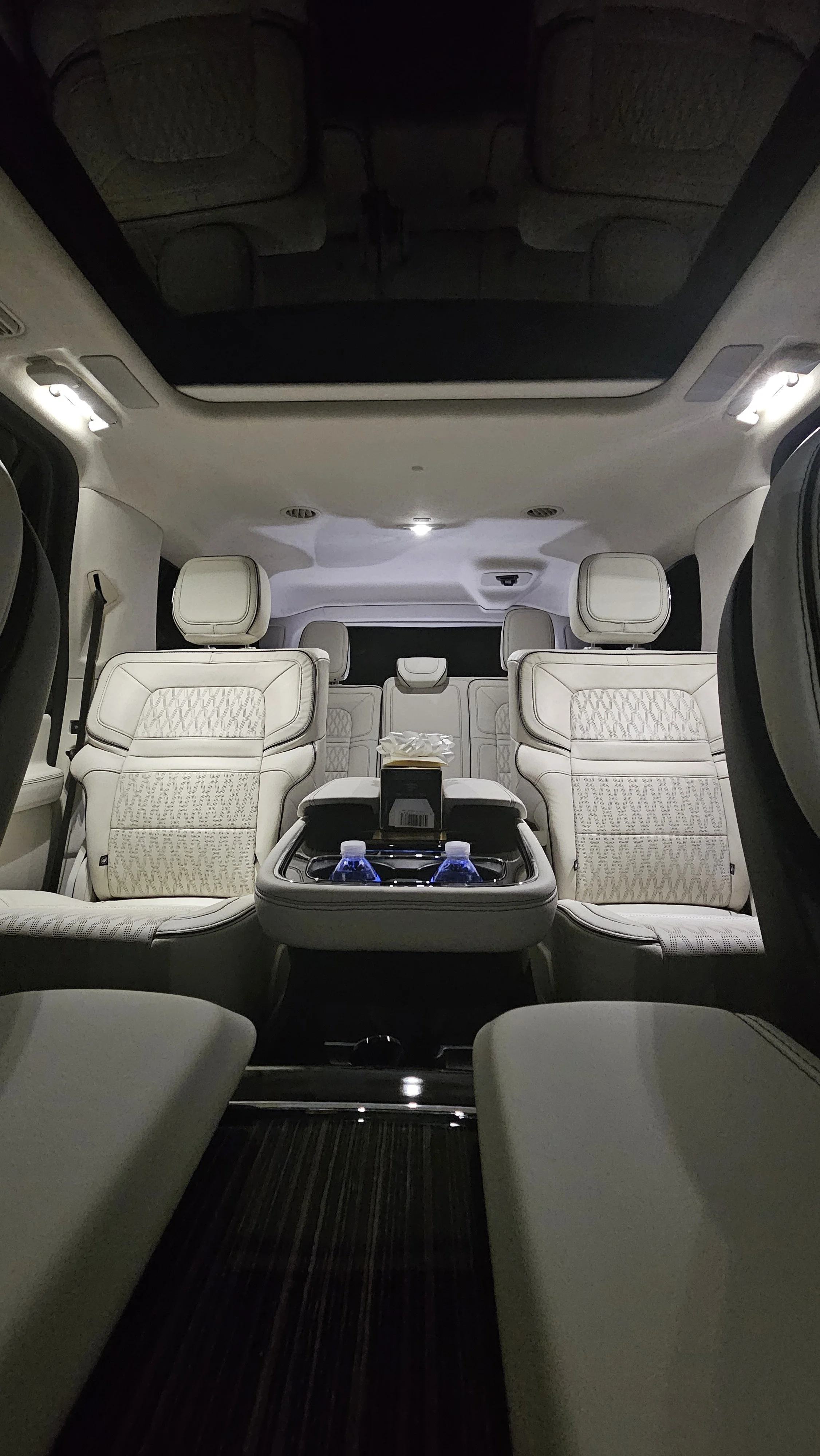 Interior of a luxury vehicle showing beige leather seats, a center console with two water bottles, tissue box, and overhead lighting, with a panoramic sunroof.