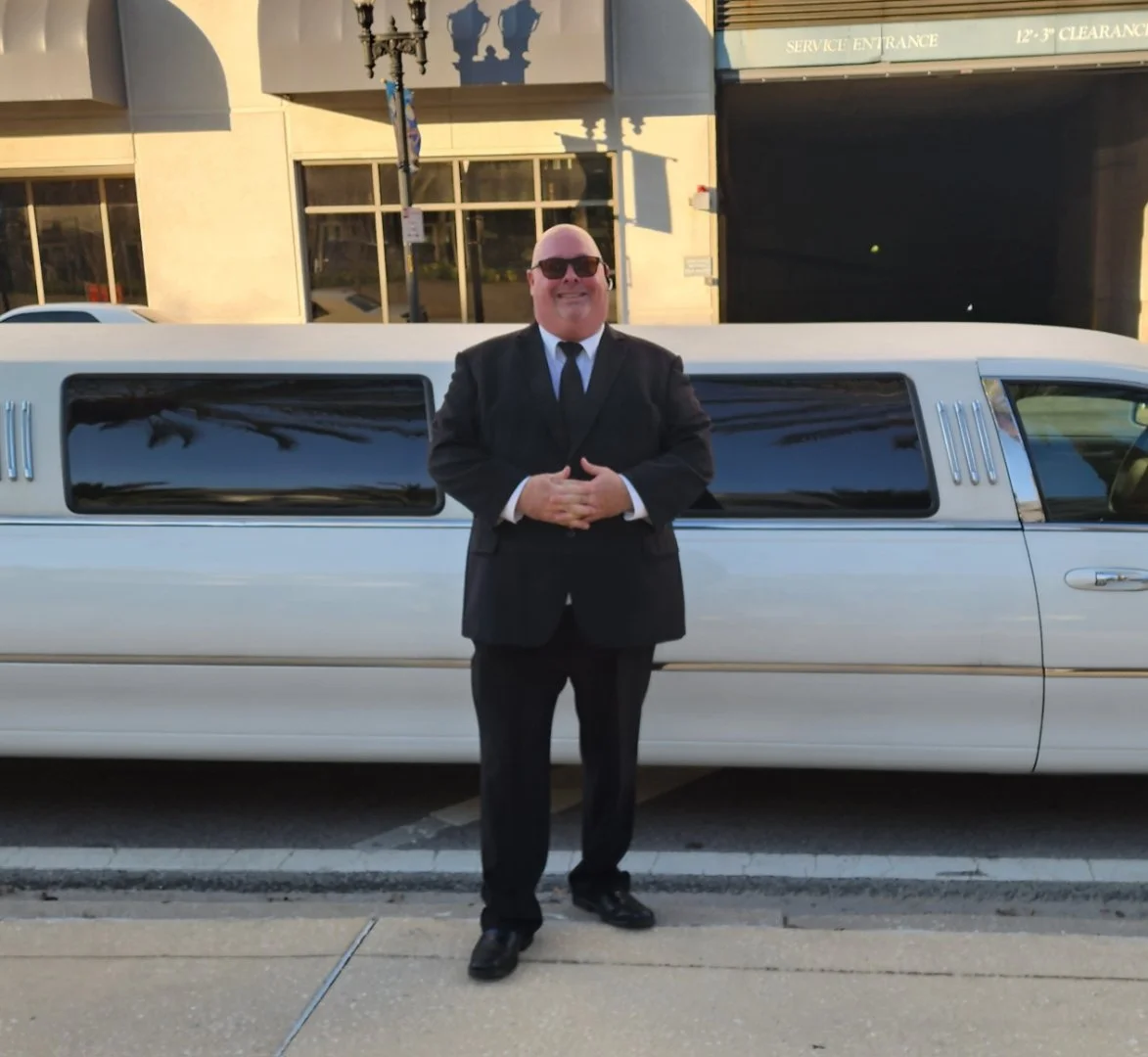 A man in a black suit, white shirt, black tie, and sunglasses standing in front of a white limousine with large tinted windows, on a city street during the day.