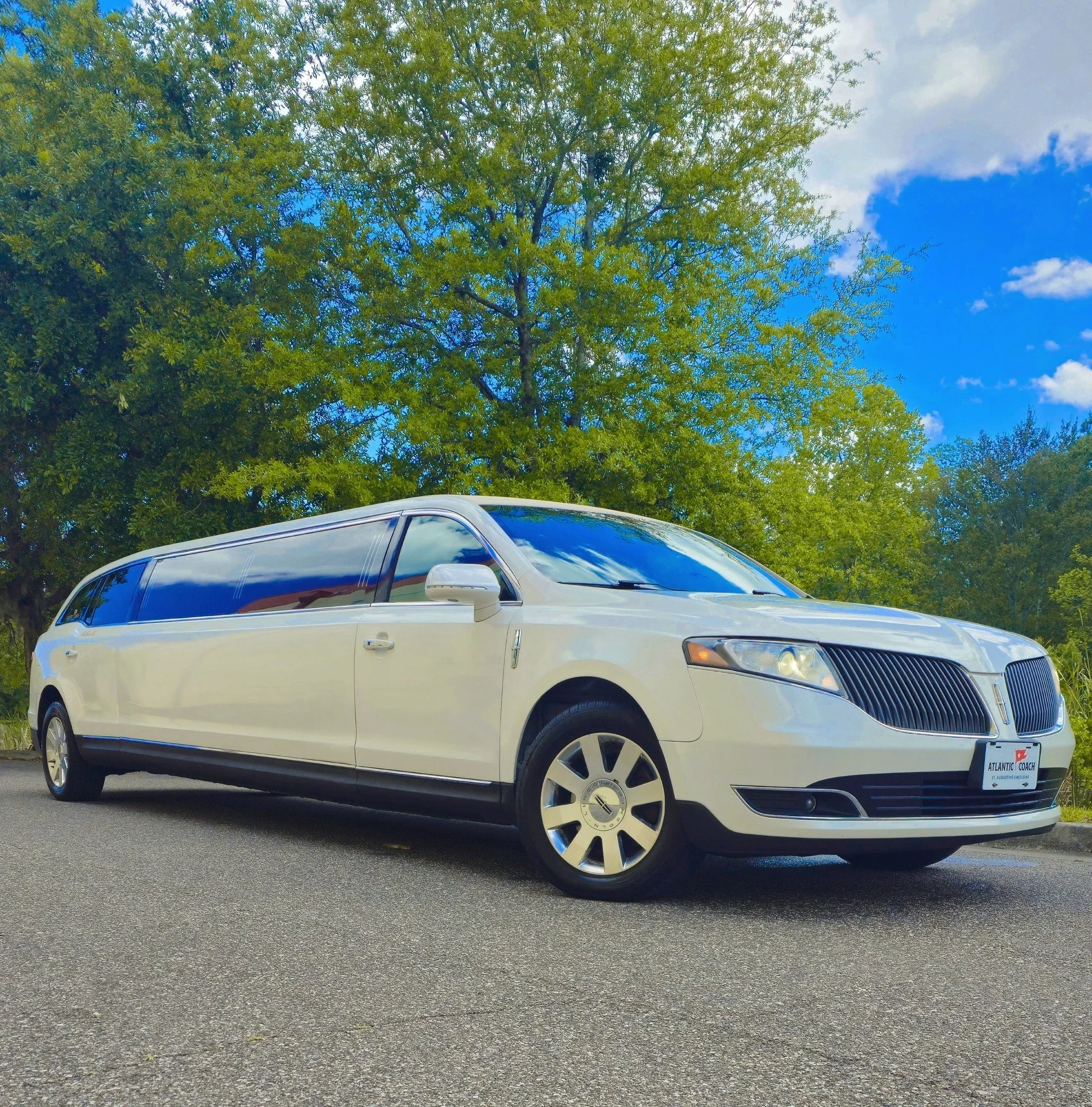 A white limousine parked on the side of a paved road with lush green trees and a bright blue sky in the background.