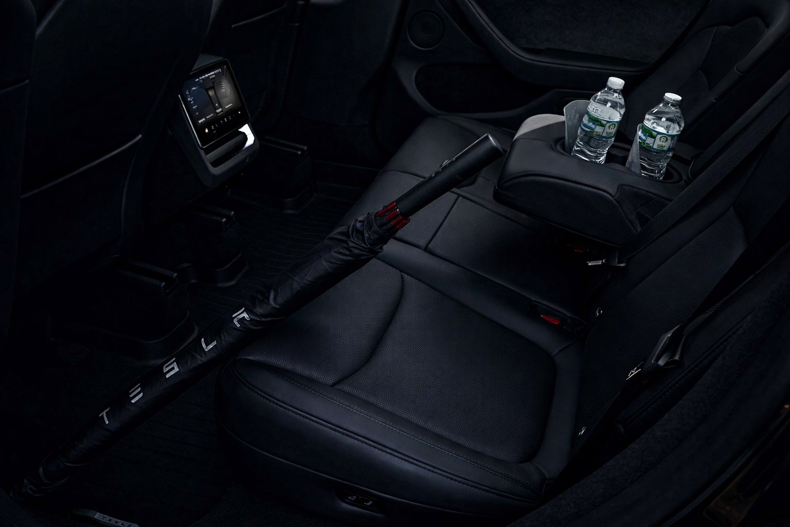 Back seat of a Tesla vehicle with a steering yoke, a central console with two water bottles, and a touchscreen display on the front dashboard.