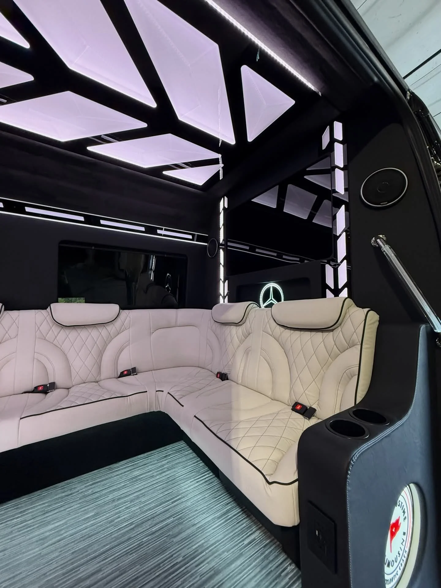 Interior of a vehicle with a beige quilted L-shaped leather sofa, black accents, and a Mercedes-Benz logo on the wall. The ceiling has illuminated geometric panels, and there are cup holders and a speaker on the side panel.