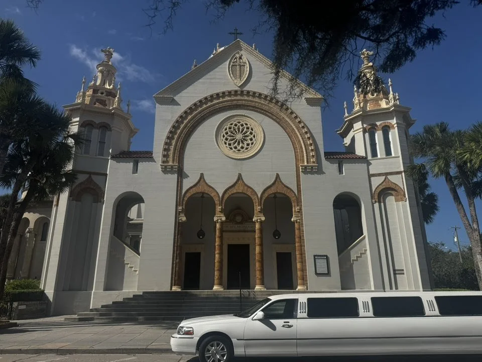 A large white church with ornate architecture, two towers on either side, and a decorative circular window above the entrance. A white limousine is parked in front. Tall palm trees are nearby against a blue sky.