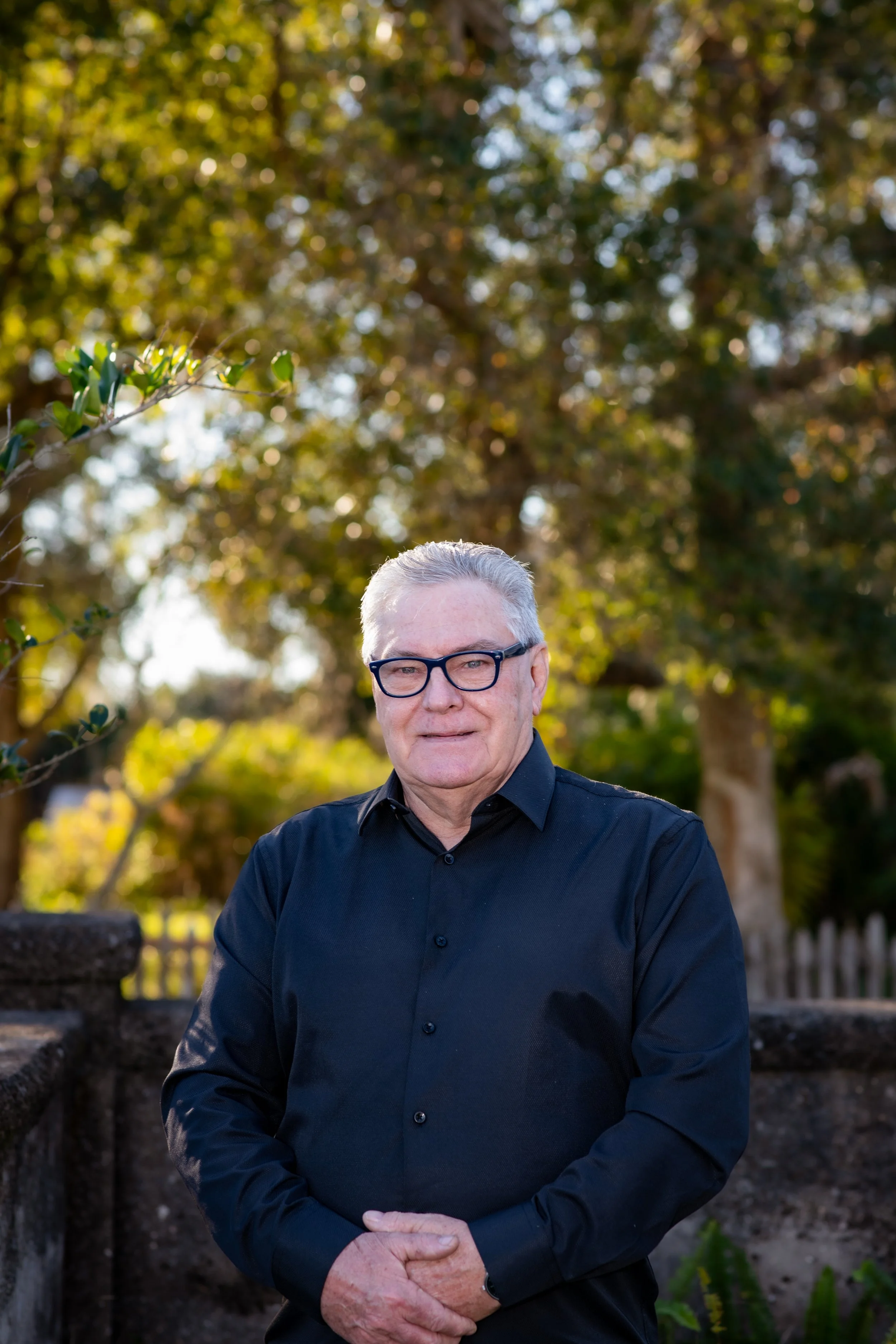 An older man with gray hair, glasses, and light skin wearing a black button-up shirt standing outdoors in front of trees during daytime.