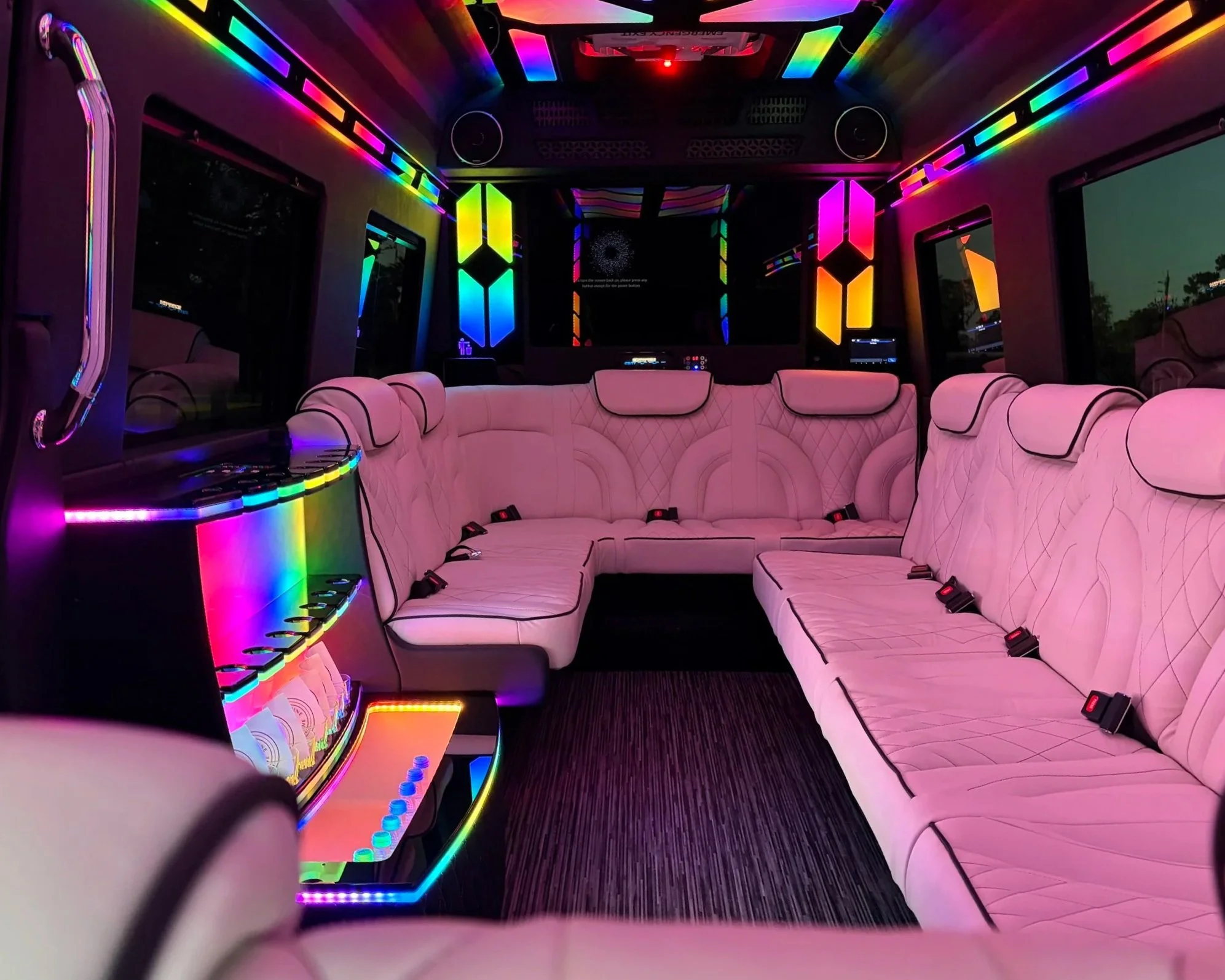 Interior of a limo van decorated with colorful LED lights, adorned with pink quilted seats, windows, and some screens or displays on the walls.
