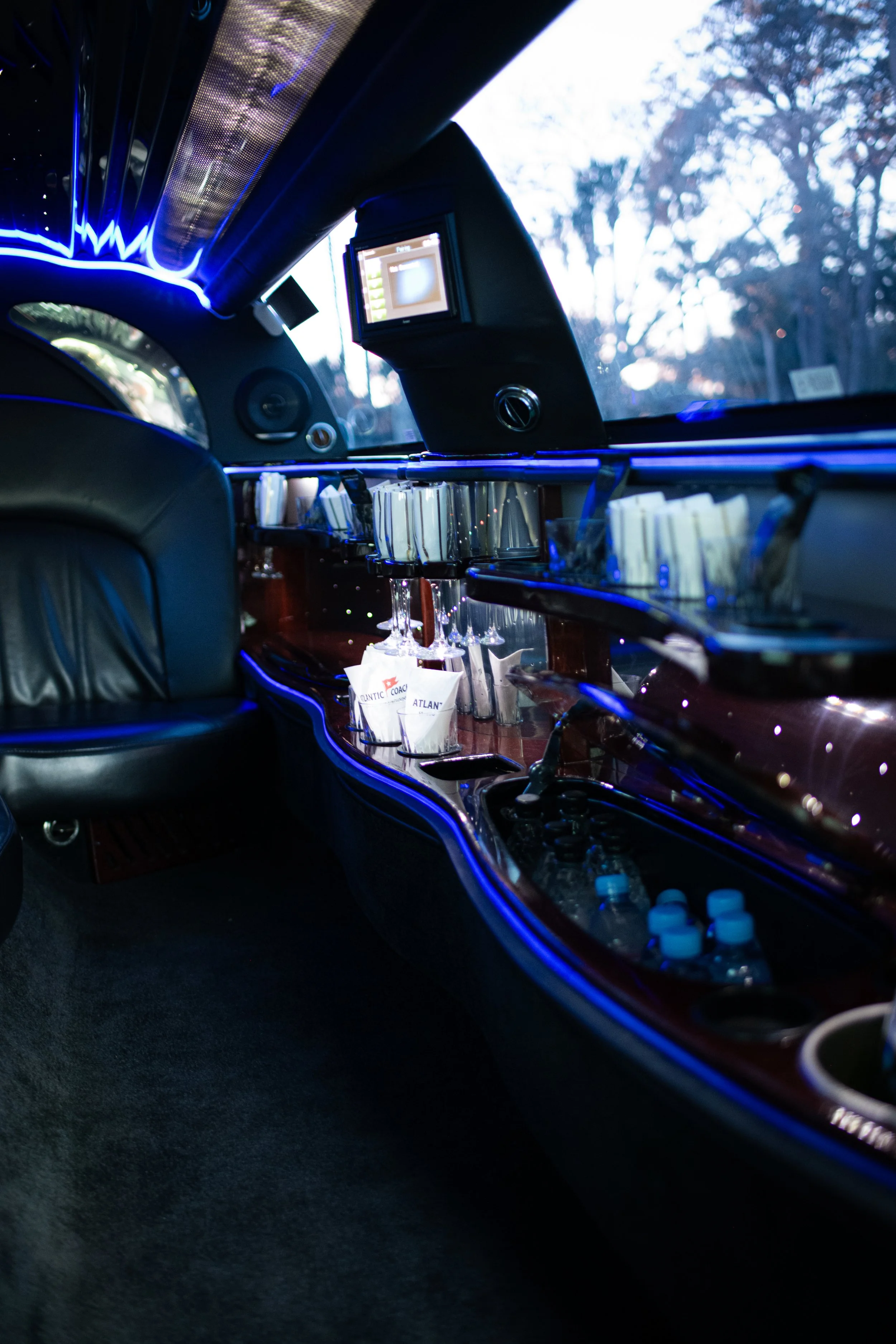 Interior of a luxury limousine with a bar area, illuminated by blue LED lights, featuring glassware, bottled water, and a TV screen.