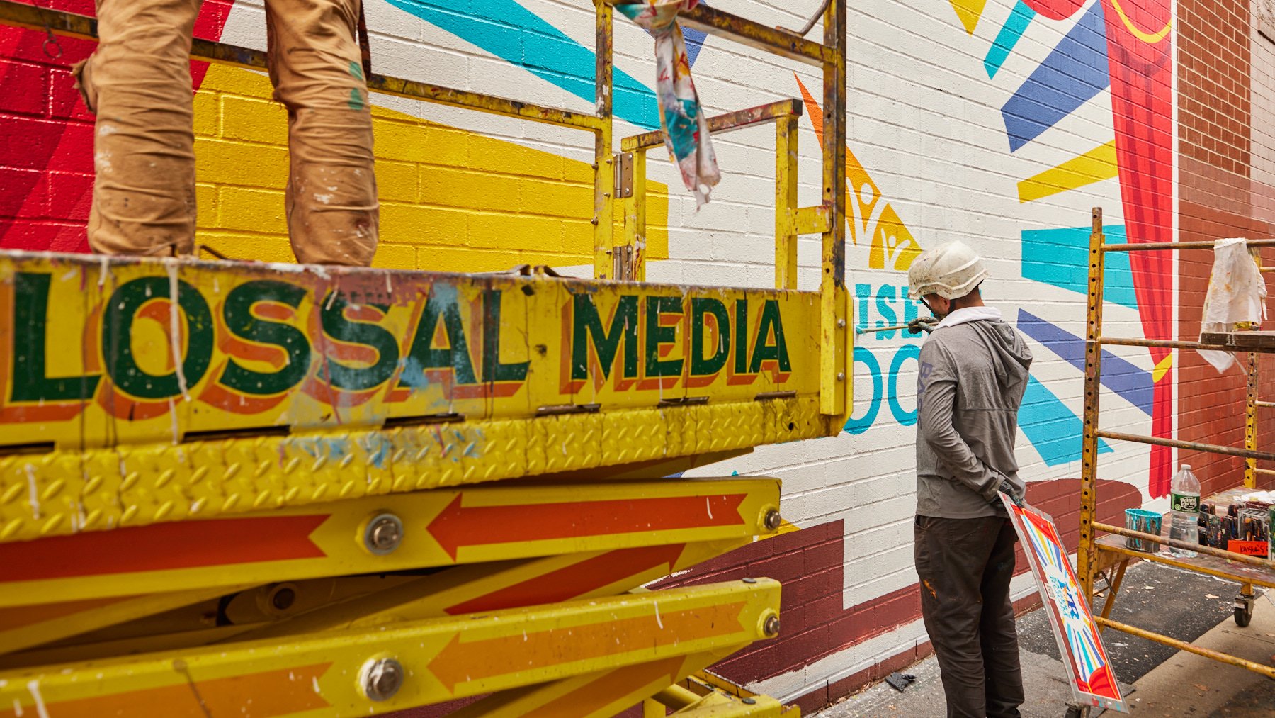 Colossal Media painters working on the Kaiser's Room mural in Williamsburg, Brooklyn. The image includes the legs of a painter standing a yellow scissor lift that reads Colossal Media with a painter to the right standing on the ground.