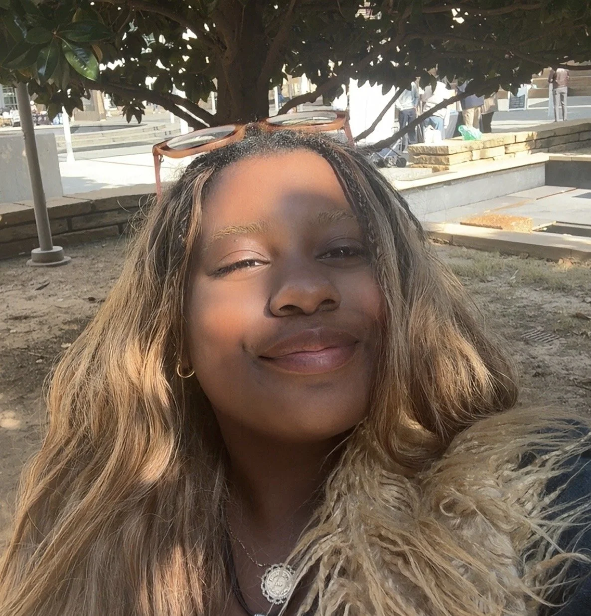 A woman with wavy, reddish-brown hair and black glasses smiling, wearing an orange sleeveless top and a delicate gold necklace with a teardrop pendant, indoors with a neutral background.