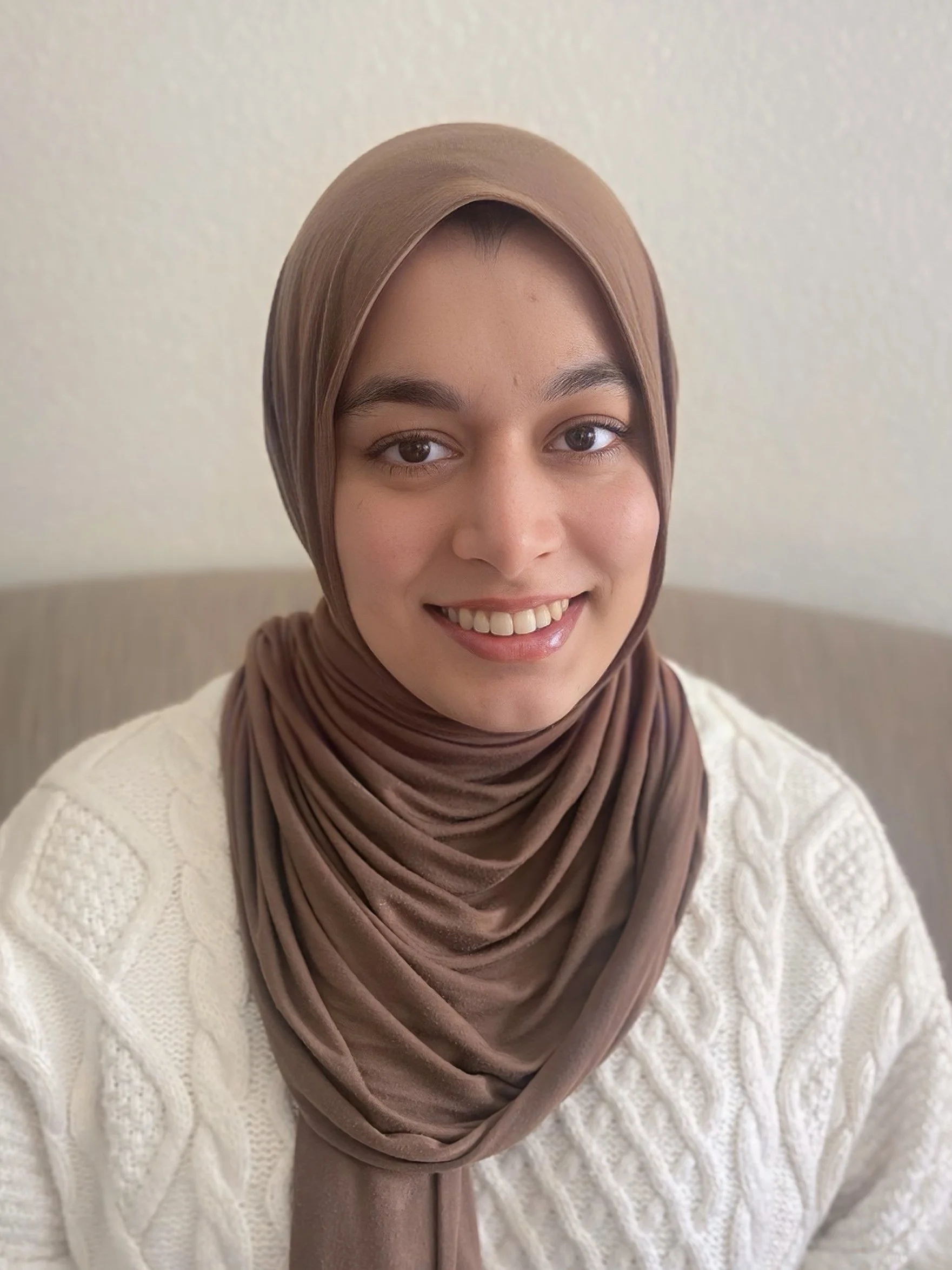 Young woman with long, dark wavy hair smiling outdoors with wooden structure and blue sky in background.