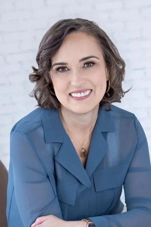 A woman with long dark brown wavy hair, smiling, wearing a black top with lace detail, and a ring on her left hand, sitting against a plain light background.
