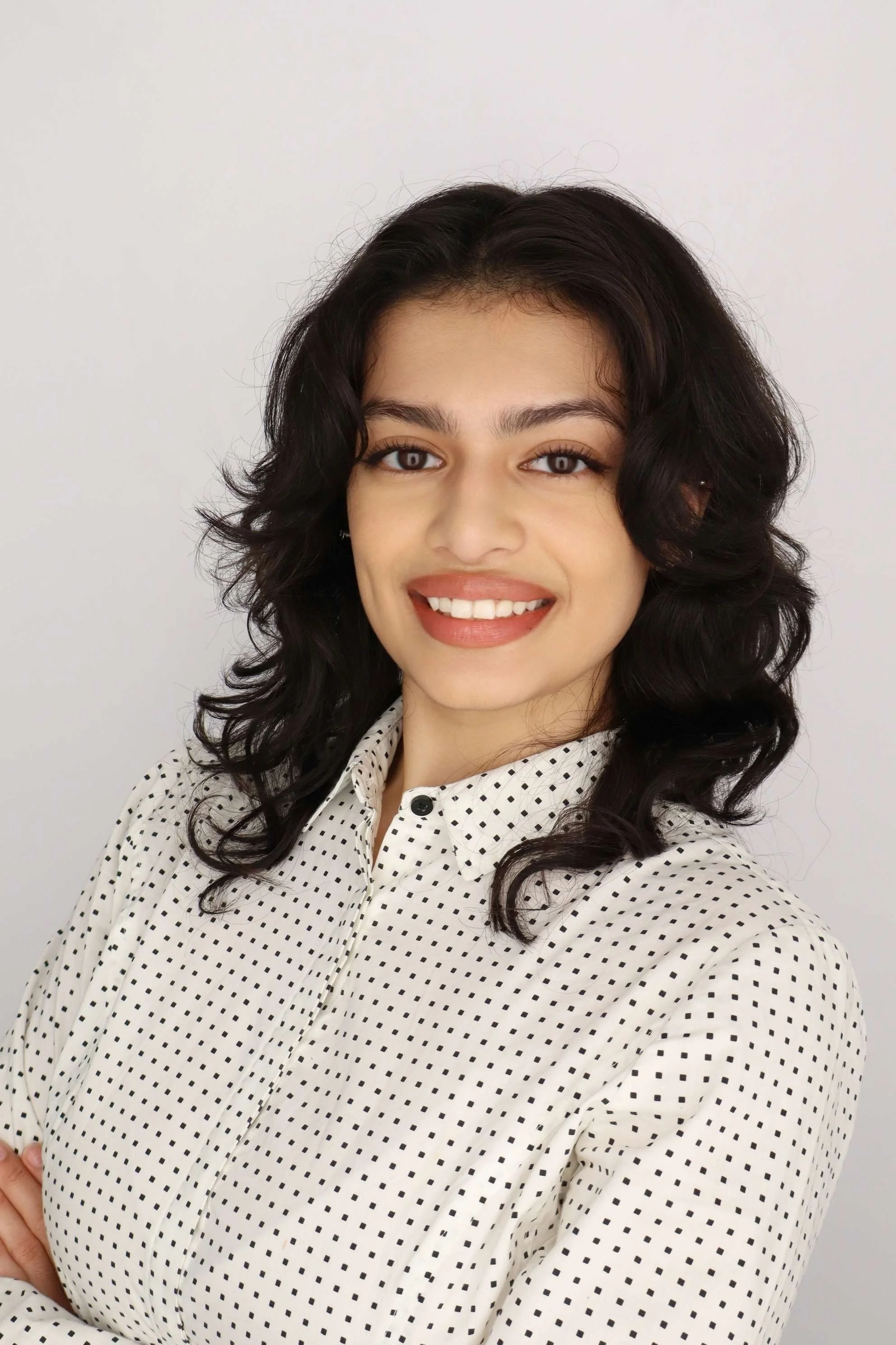 A young woman outdoors with long black hair, wearing glasses, a blue and white striped blouse, and a white top underneath, standing on a grassy field.