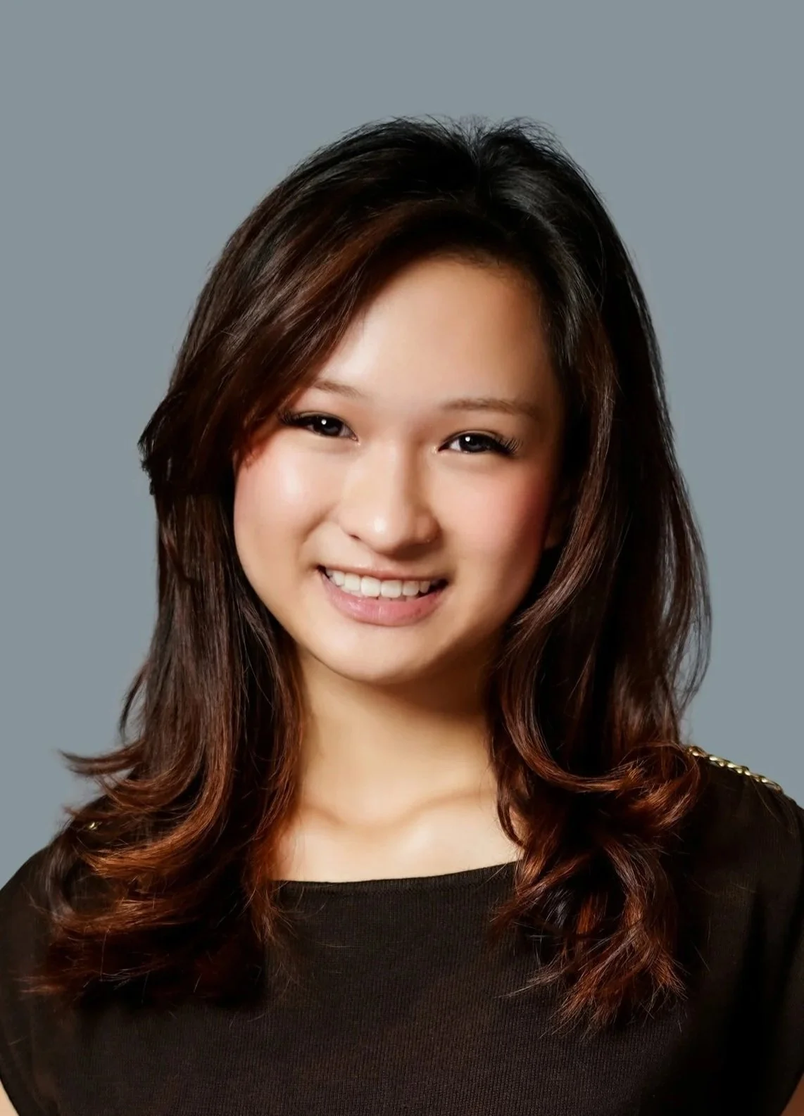 Young woman with long, dark wavy hair smiling outdoors with wooden structure and blue sky in background.