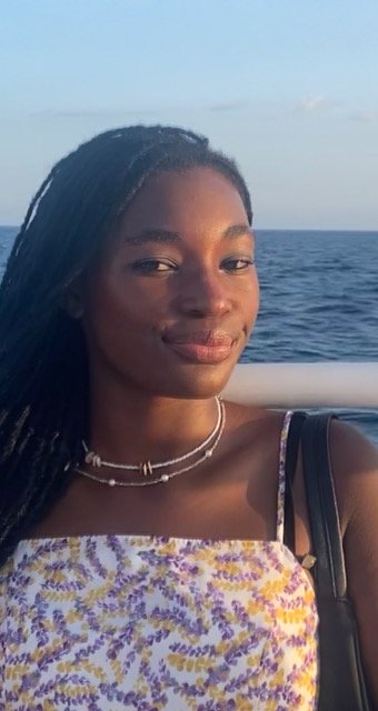 A young woman with long brown hair smiling at the camera, wearing a light blue top and a gold necklace, against a plain light background.