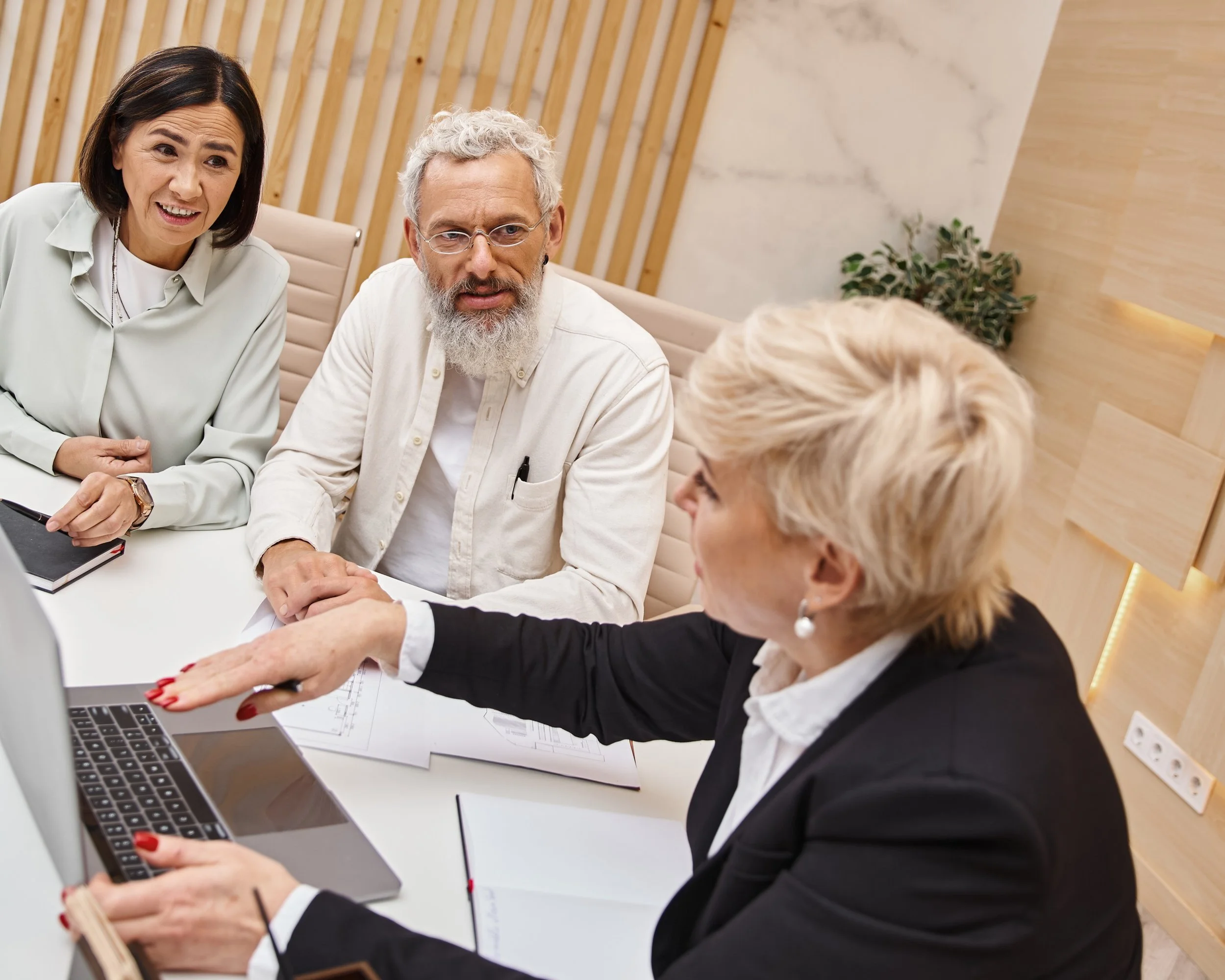 Three business professionals in a meeting room discussing work, with one woman pointing at a laptop screen.