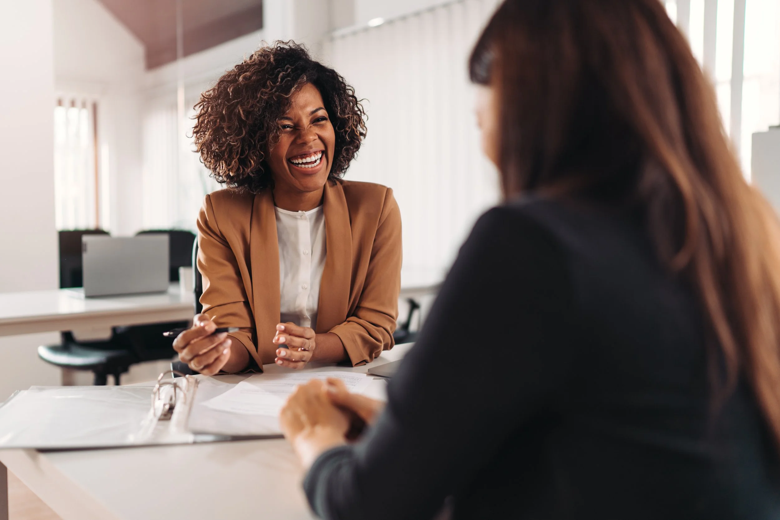 Two women in an office, one with curly hair wearing a brown blazer, the other with straight hair in a dark blazer, engaged in a conversation, smiling, with papers on the table.