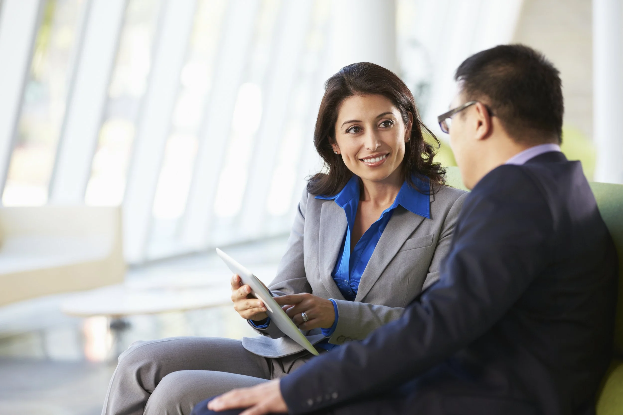 A woman with dark hair wearing a gray blazer and blue blouse having a conversation with a man in glasses and a dark suit at a modern office lounge.