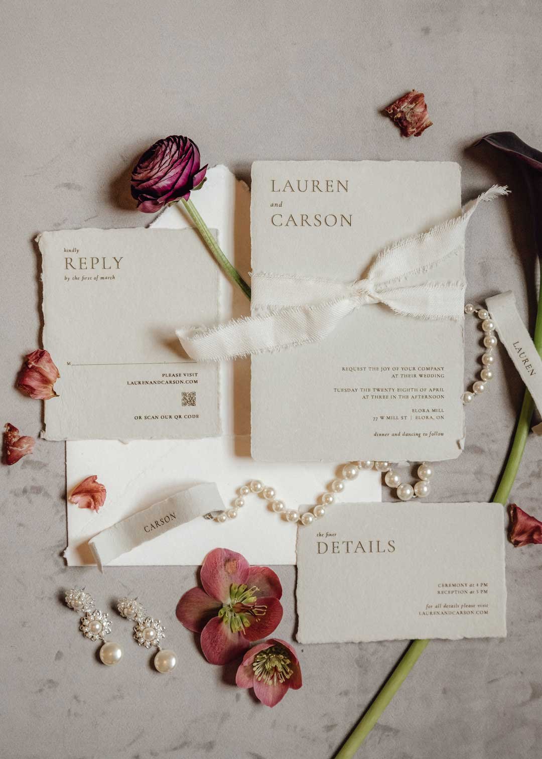 Wedding invitation suite with floral decorations and pearl jewelry, featuring cream-colored paper with black text, pink ranunculus flowers, and scattered rose petals.