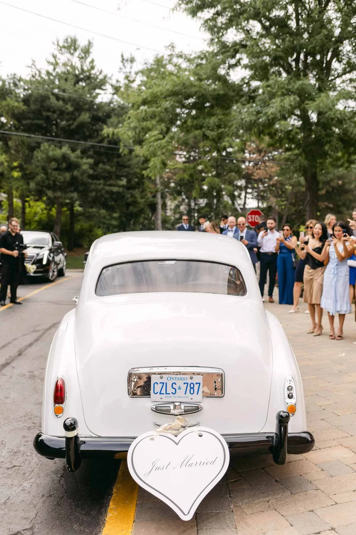A white vintage car decorated for a wedding, with a heart-shaped sign that reads "Just Married" attached to the back. The car is parked on a street, and a group of people are gathered on the sidewalk, celebrating and taking photos.