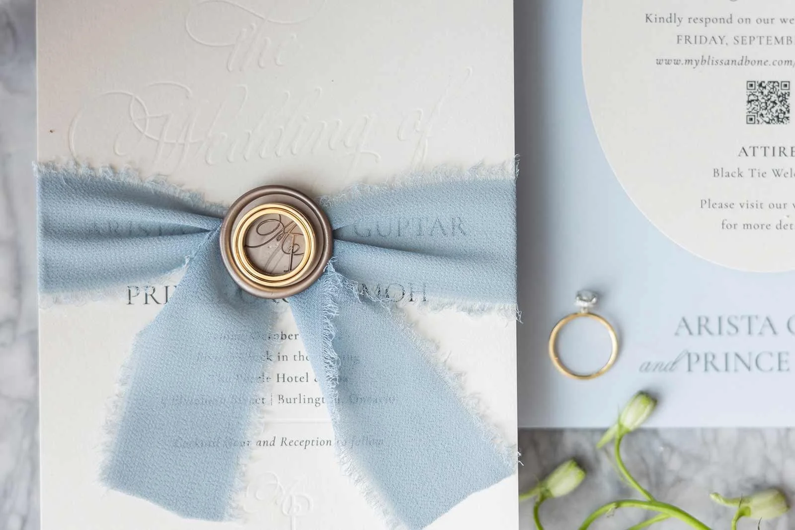 Wedding invitation with a light blue ribbon and gold ring embellishments. The invitation has embossed text and a QR code, surrounded by small white flowers.