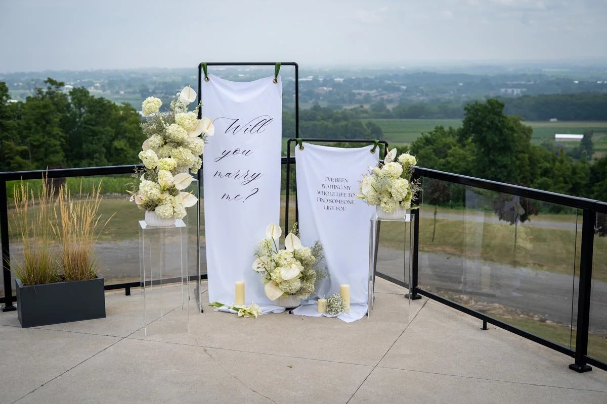 Wedding ceremony backdrop with white fabric banners containing handwritten and printed text, surrounded by white floral arrangements, candles, and potted plants on a terrace overlooking a rural landscape.
