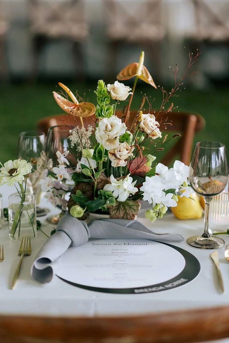 A table setting with a floral centerpiece, wine glasses, and a menu titled "Savour the Moment," with outdoor seating in the background.