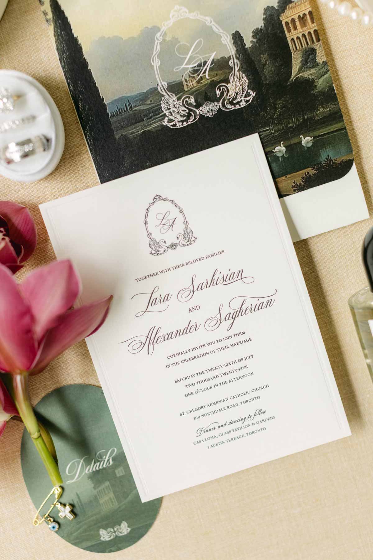 Wedding invitation card with elegant script, floral design, and monogram, placed on a beige tablecloth next to pink flowers, a photo of a landscape with swans, and jewelry.
