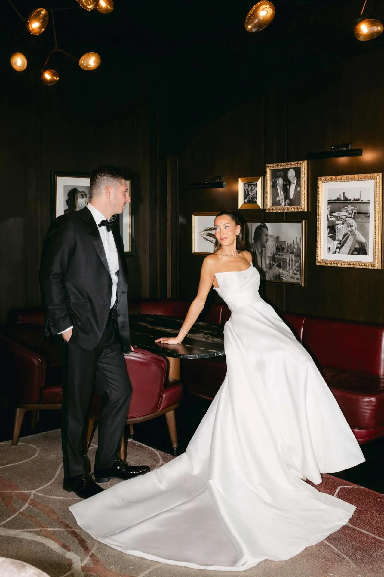A bride in a white wedding dress and a groom in a black tuxedo standing in a dimly lit room with framed black and white photographs on the wall.