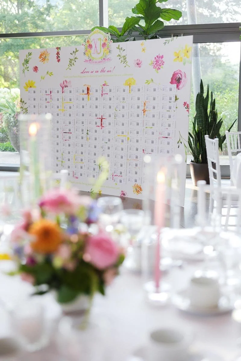A seating chart with floral decorations and the phrase "Love is in the air!" at the top, set up in a bright room with large windows and visible greenery outside. Blurred flowers and tableware are in the foreground.